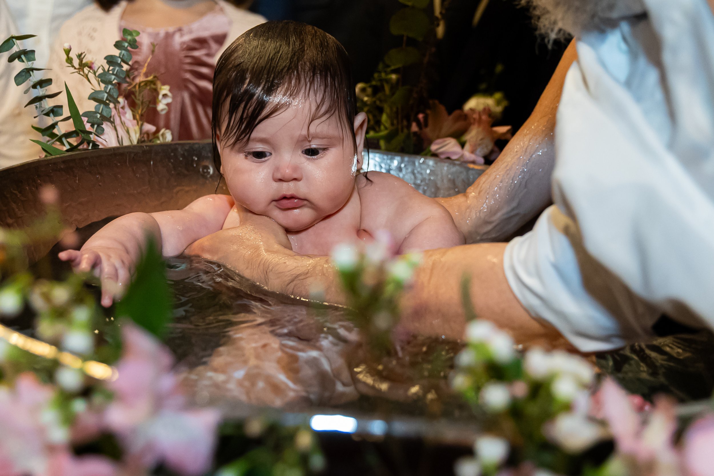 A baby being baptized in a small basin by a person in a white shirt, with greenery and flowers in the foreground.