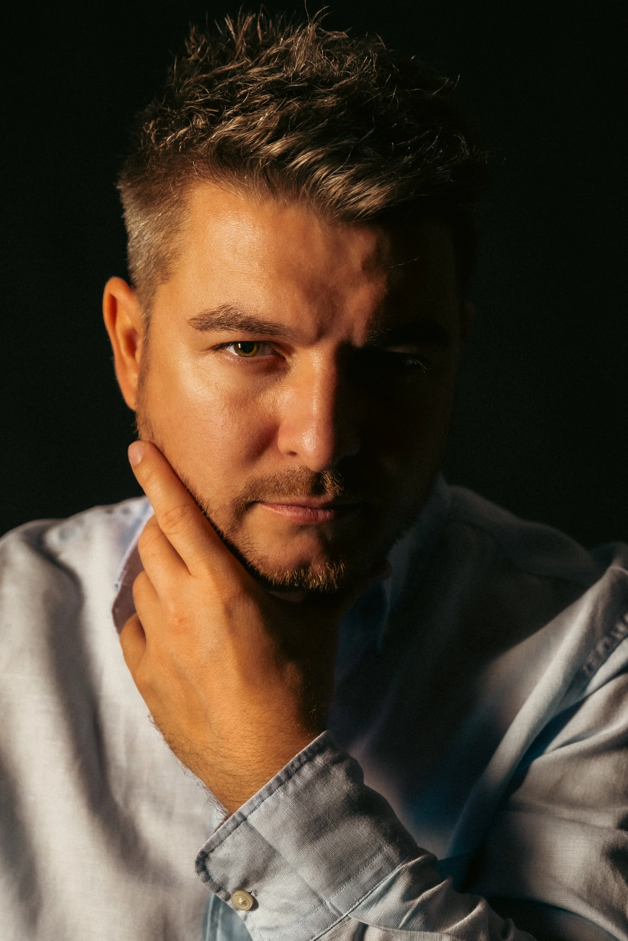 Close-up of a young man with styled short hair, light beard, and light brown eyes, wearing a light-colored, checkered shirt, and posing with his hand near his chin against a dark background.