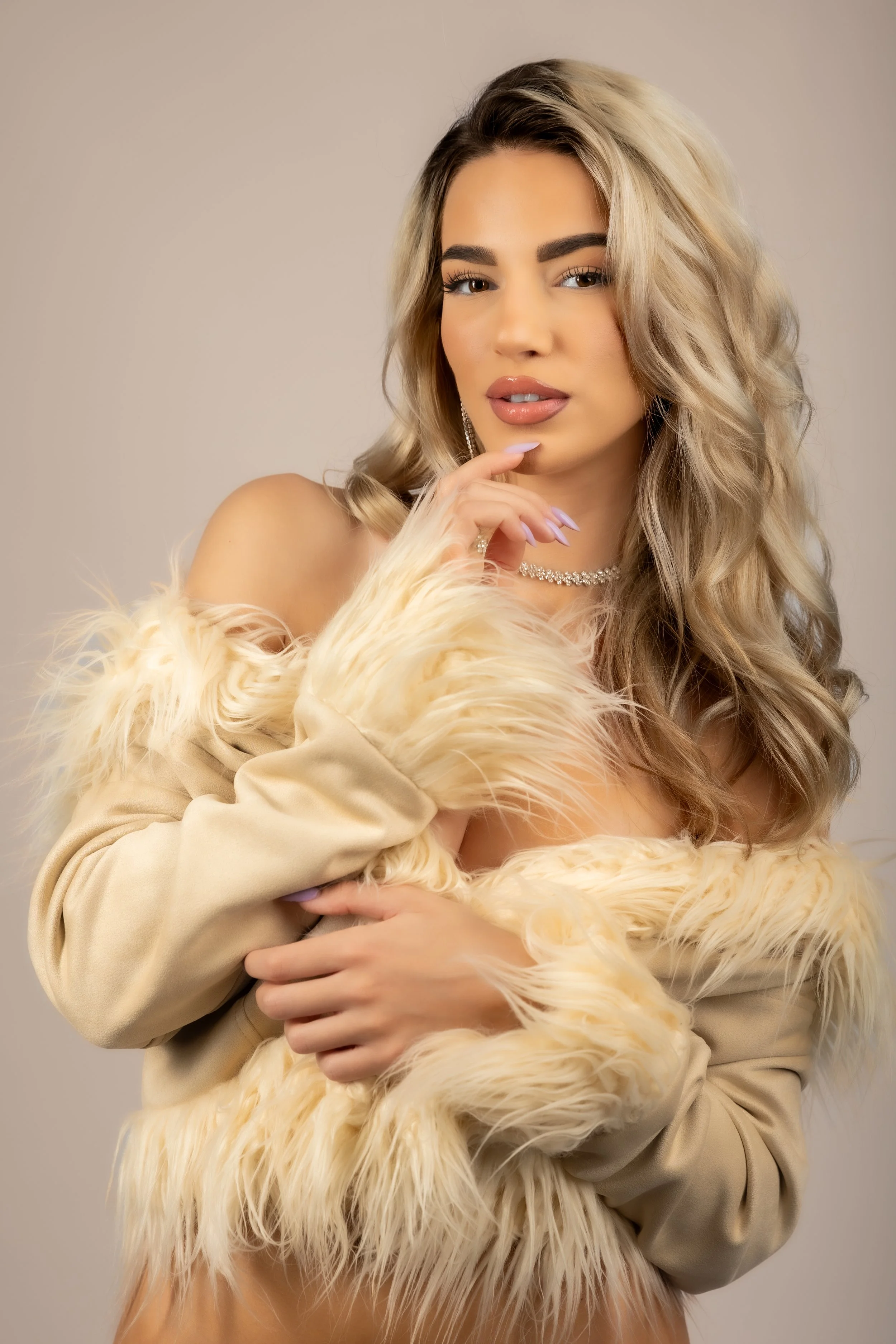 A woman with blonde wavy hair wearing a beige fur coat and jewelry, striking a pose against a plain background.