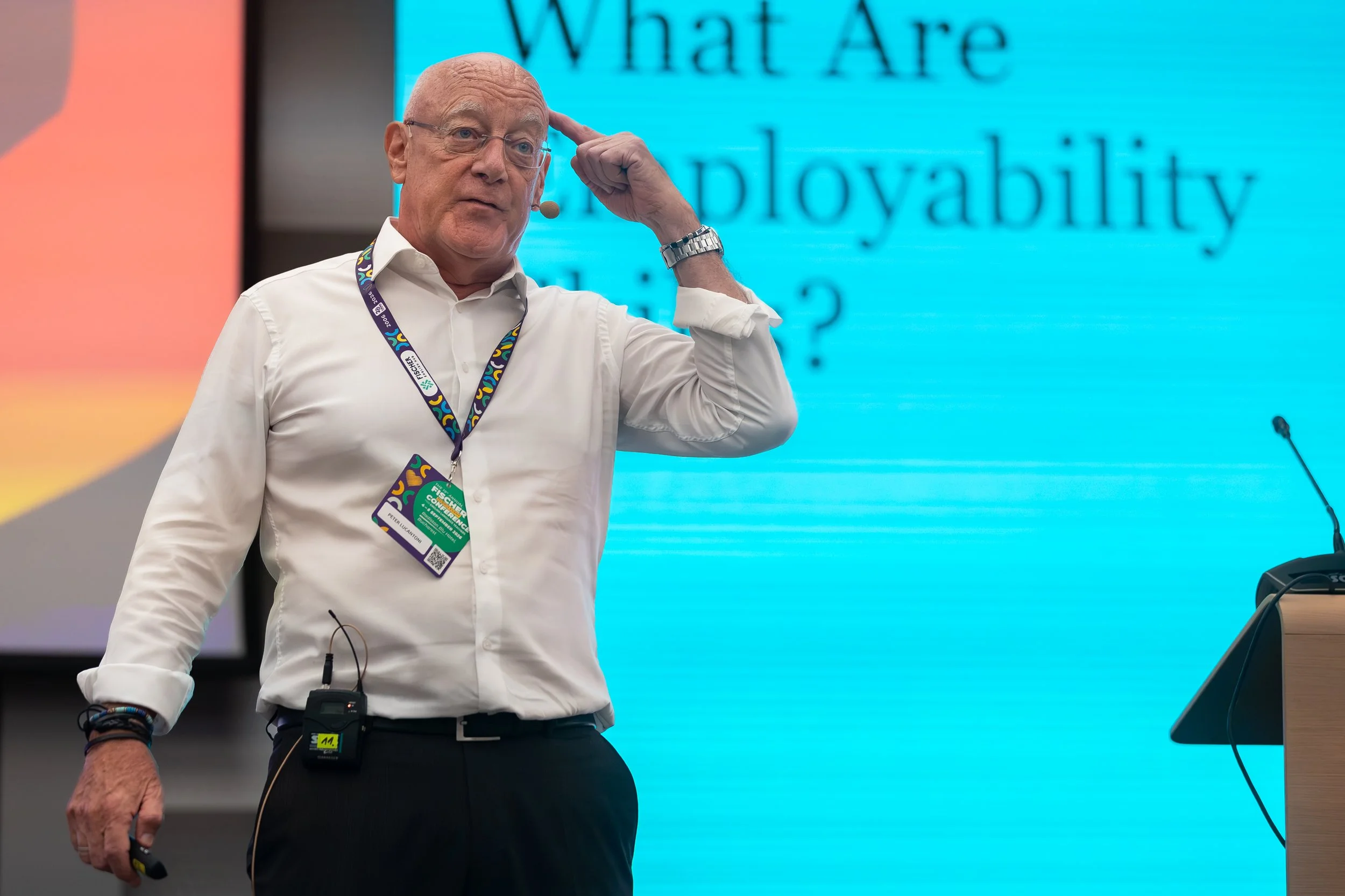 A man with glasses and a white shirt is standing in front of a blue screen that has the words 'What Are' and 'lability' visible. He has a watch, microphone, and conference badge on his neck and is adjusting his glasses while speaking at a conference.