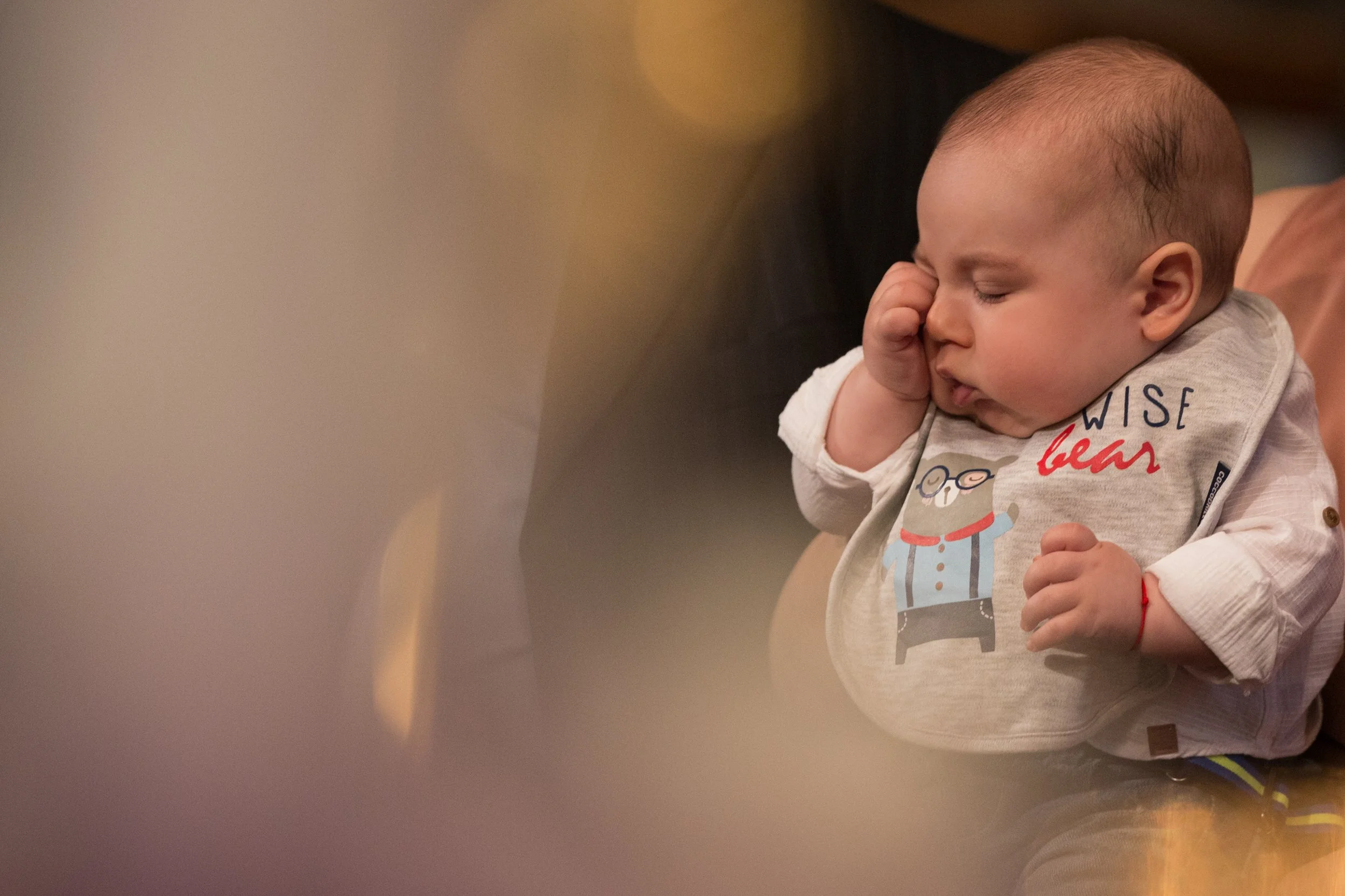 A young child with a shaved head, wearing a gray shirt with colorful graphic, appears to be resting with eyes closed and hand on face, sitting indoors.