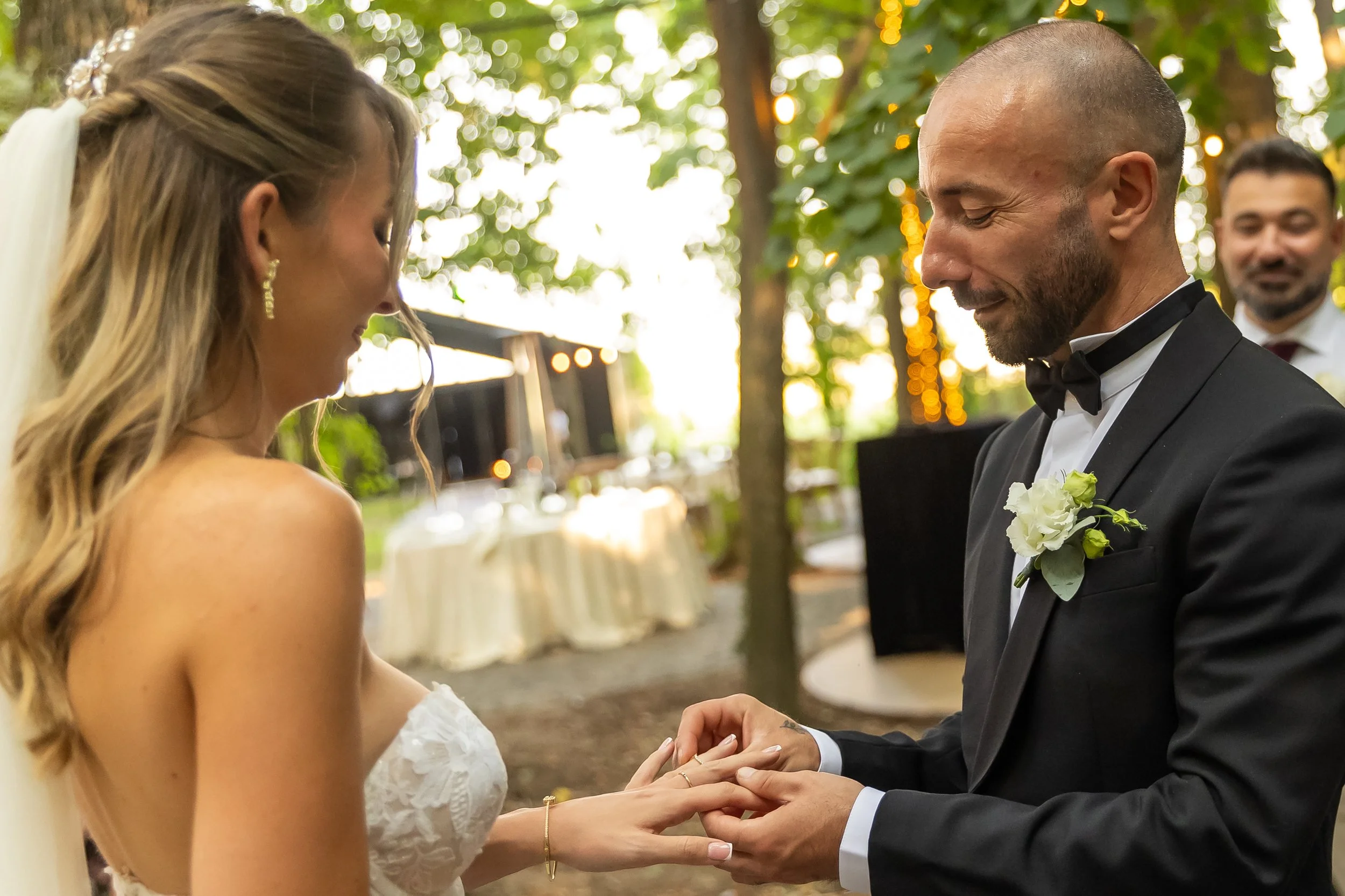 A bride and groom holding hands during their wedding ceremony outdoors, with a person smiling in the background, surrounded by trees and warm lighting.