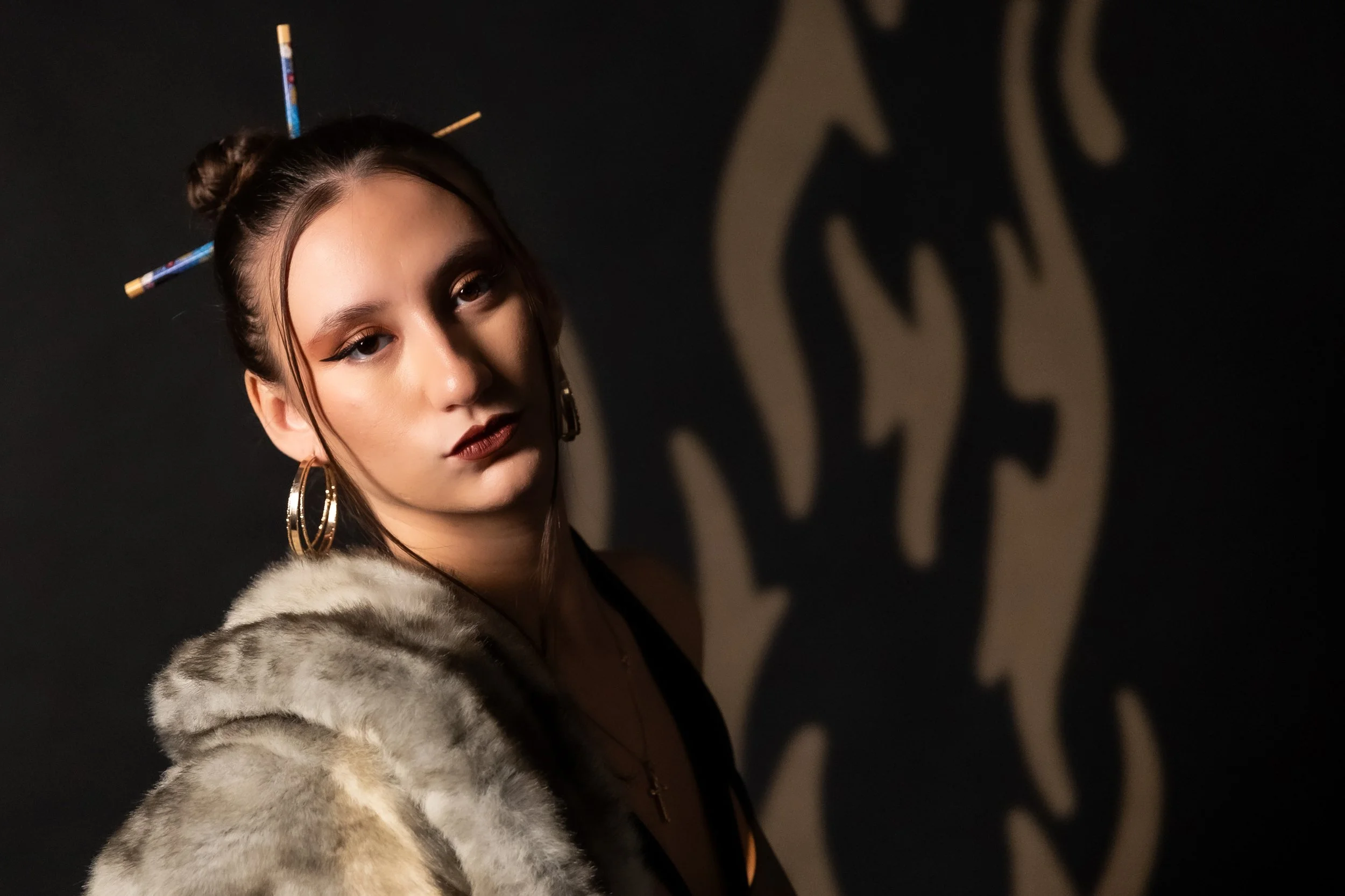 A young woman with dark hair styled in two buns, wearing large hoop earrings, makeup with winged eyeliner and dark lipstick, a faux fur coat, and colorful chopsticks in her hair, standing in front of a dark wall with a shadowed logo or design