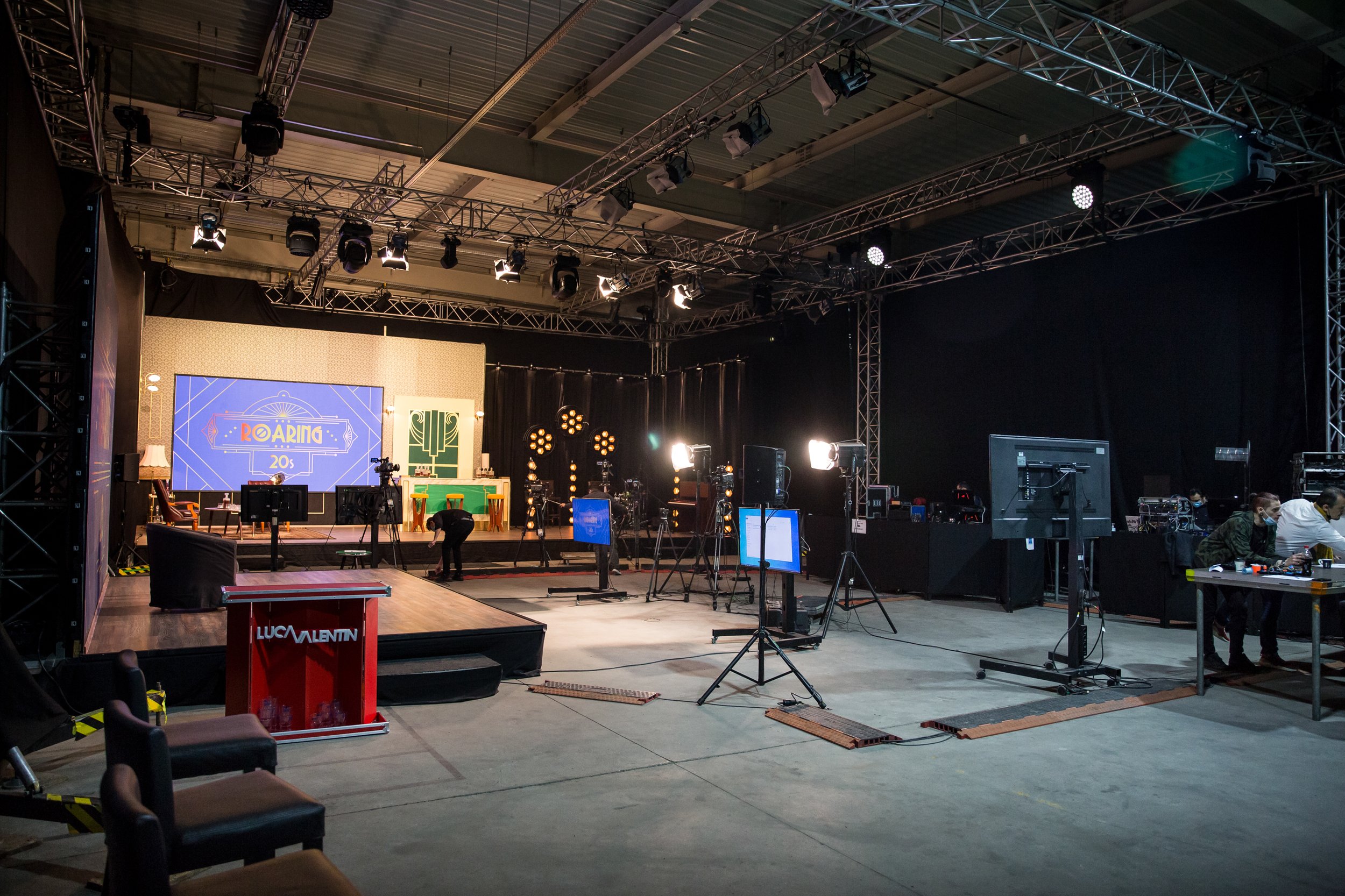 An indoor event stage setup with lighting and video equipment, a large screen displaying 'Roaring 20s' theme, and some chairs and tables around, indicating preparations for a performance or presentation.