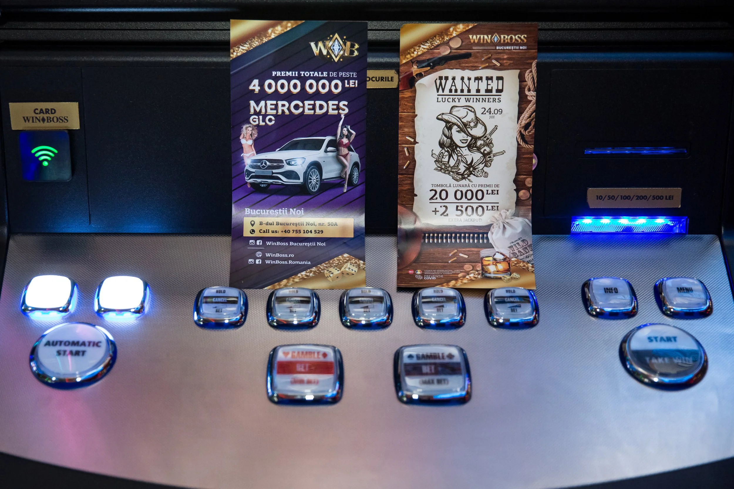 Close-up of a gaming slot machine with two promotional banners. One banner advertises winnings of over 4 million lei with a Mercedes GLC car image, and the other promotes a lottery with a woman in a cowboy hat surrounded by gambling symbols. The cont