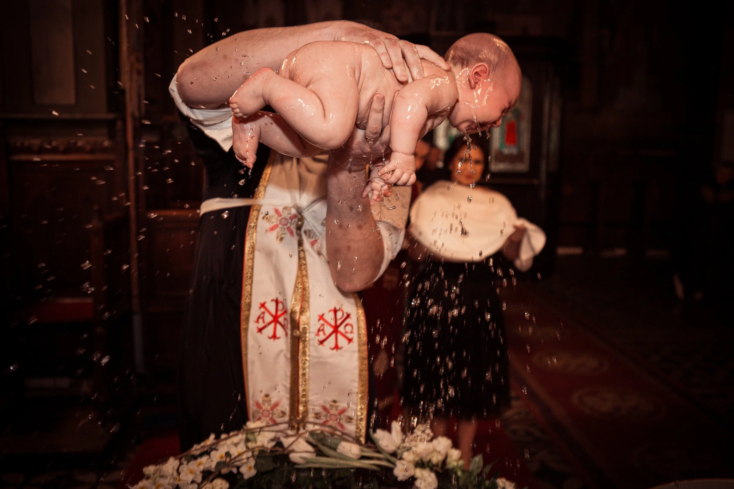 A priest holding a baby during a religious ceremony, with water splashing and flowers at the altar.