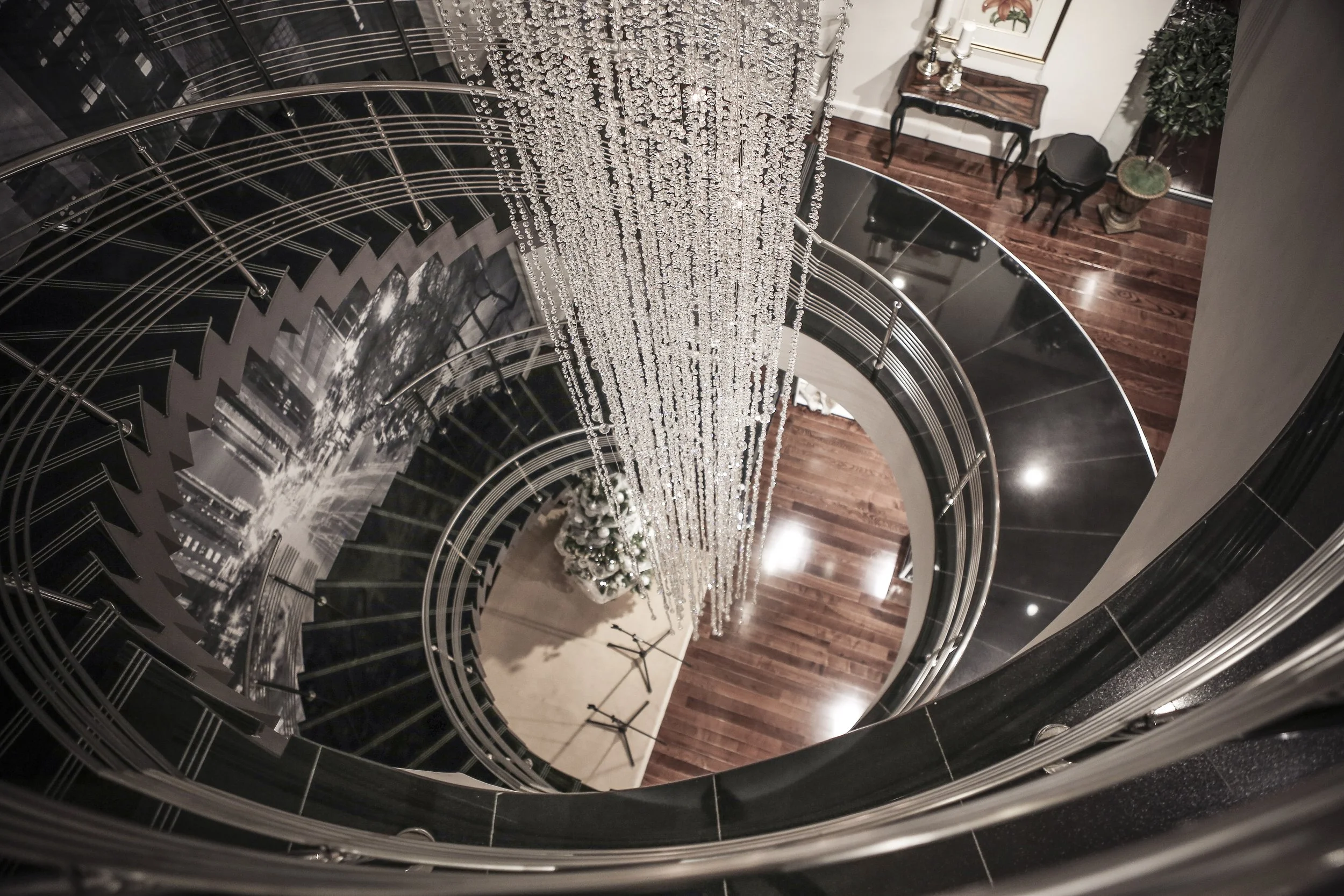 A view down a spiral staircase with black and metal railing, overlooking a wooden floor and a Christmas tree decorated with white ornaments and lights, with a chandelier hanging above.