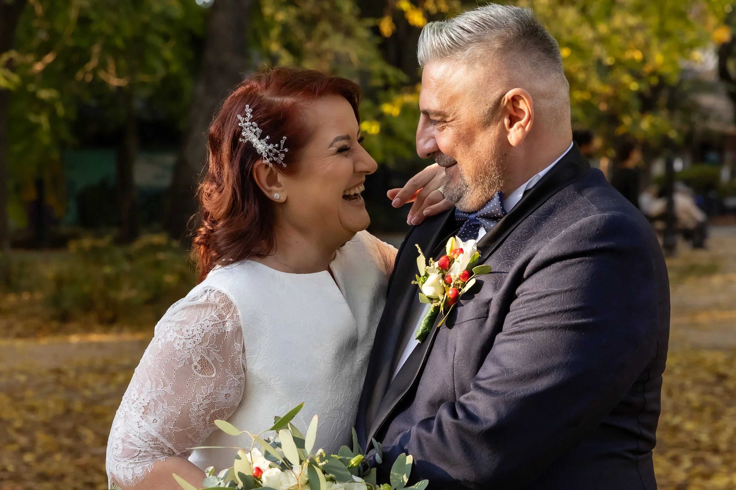 A bride and groom smiling and facing each other outdoors during a wedding, with trees and fall foliage in the background.