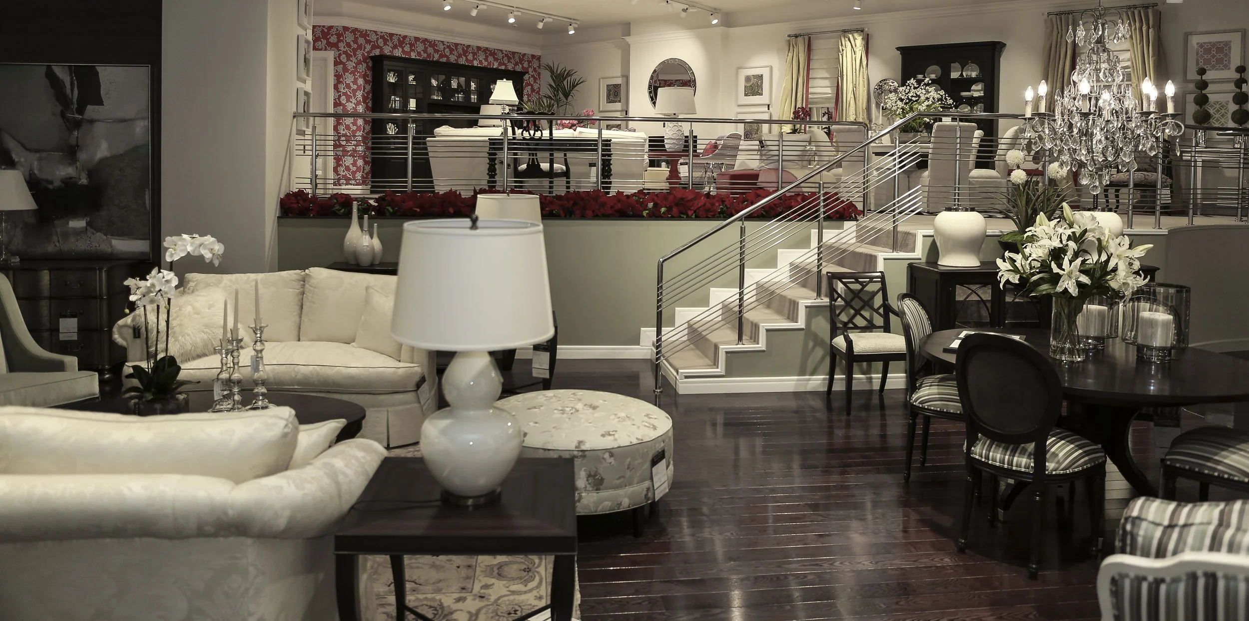 Elegant living room and dining area with white and beige furniture, floral arrangements, chandeliers, and decorative elements, with a staircase leading to an upper level.