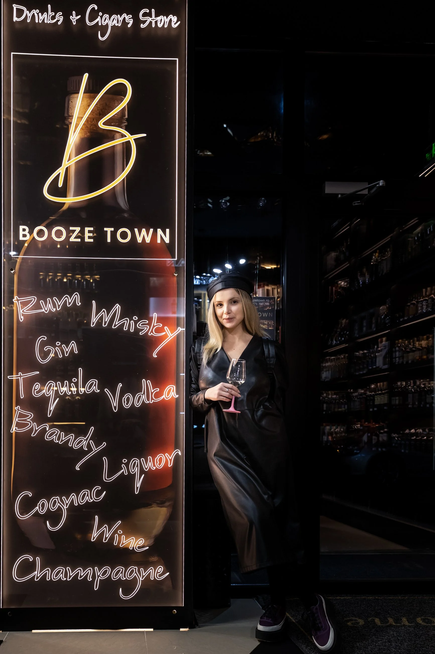 A woman with blonde hair wearing a black outfit and black cap holding a wine glass standing beside a lit sign that reads 'Drinks + Cigars Store' and 'Booze Town,' with a list of alcoholic beverages including rum, whiskey, gin, tequila, vodka, brandy,