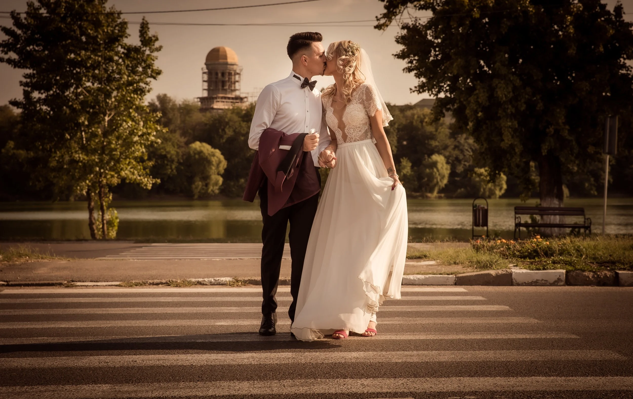 A newlywed couple in wedding attire sharing a kiss while walking across a crosswalk in a park during sunset, with trees and a body of water in the background.