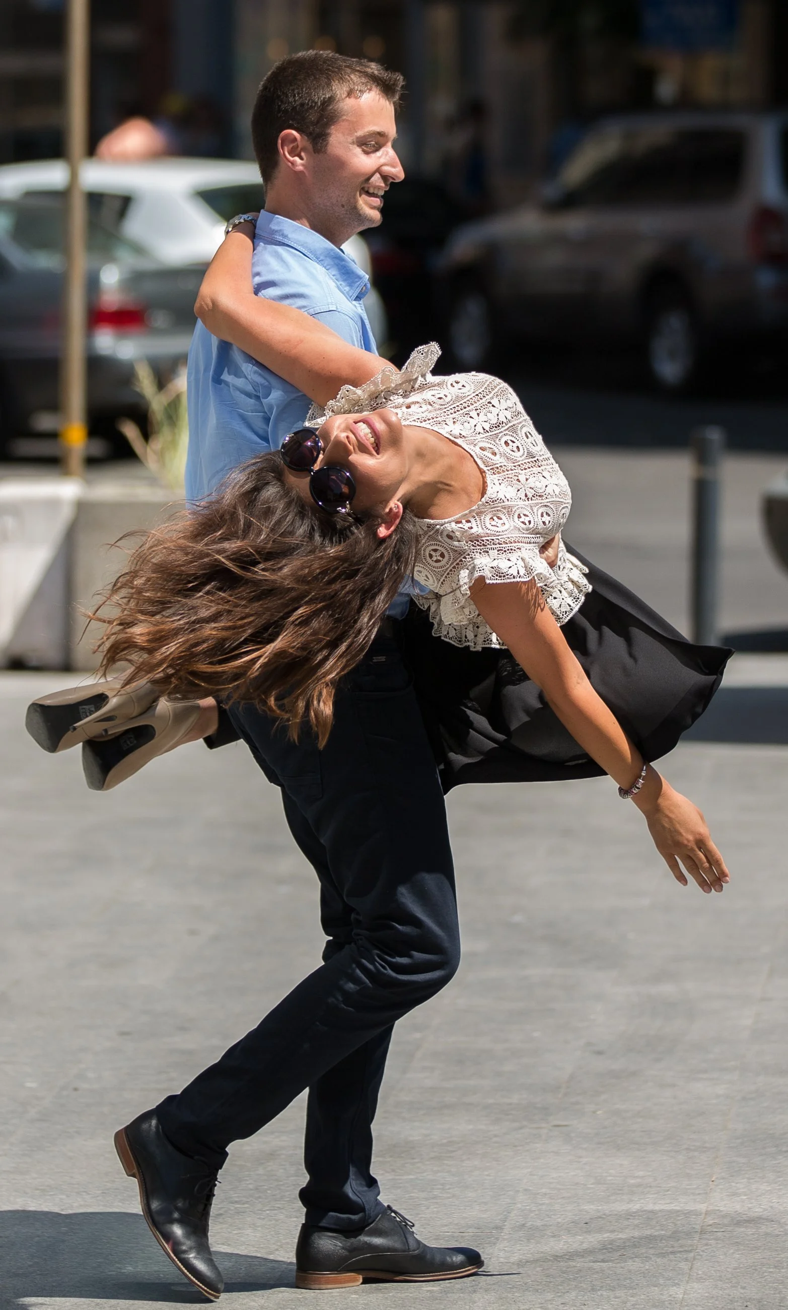 A man in a light blue shirt is holding a woman in a lace top and black skirt, who is wearing sunglasses, as she leans back with her hair flowing and a joyful expression on her face. They appear to be dancing or playful in an outdoor urban setting wit
