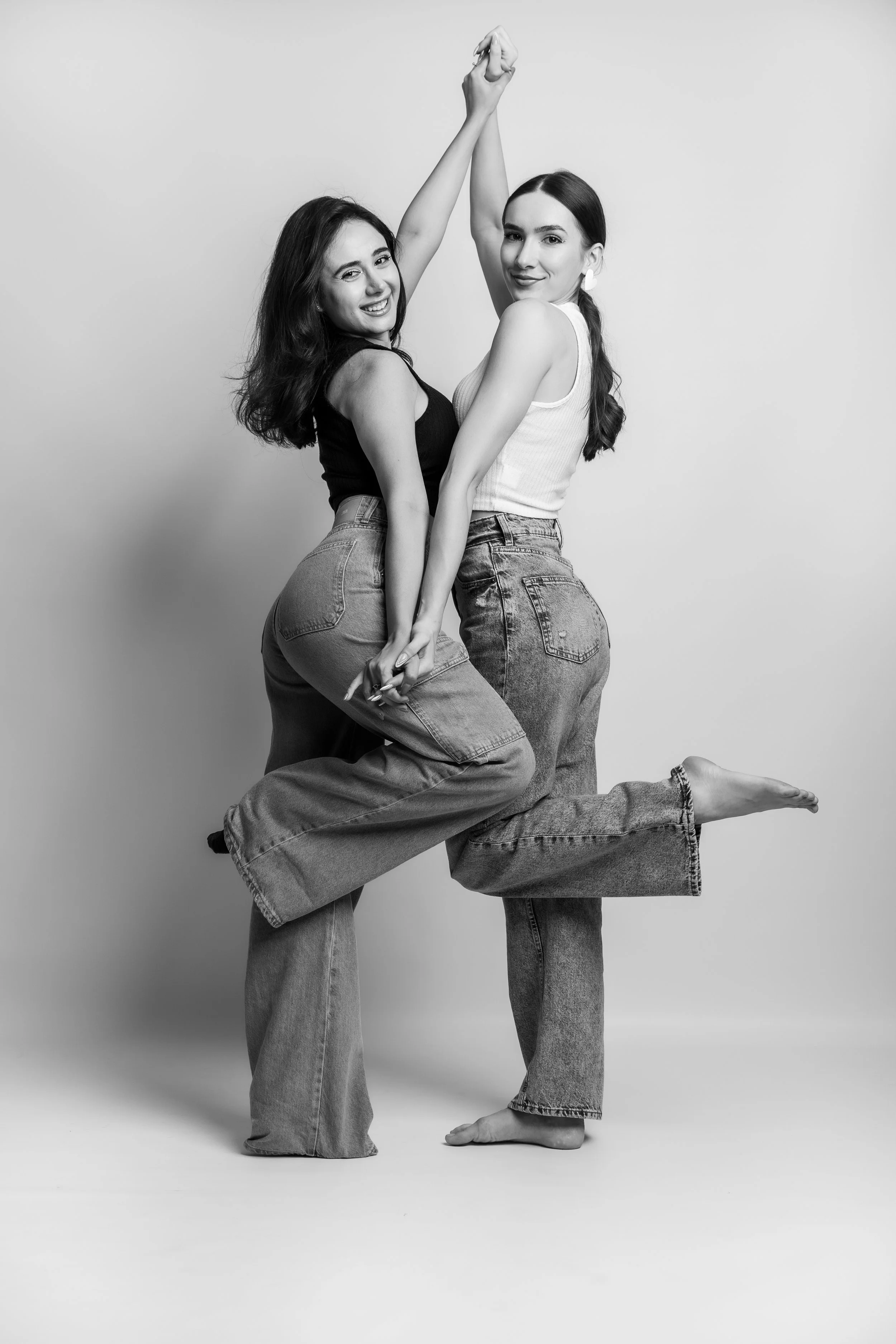 Two women pose together against a plain background, holding hands, with one woman smiling and the other with a subtle smile. They are dressed casually in tank tops and jeans, and are barefoot.