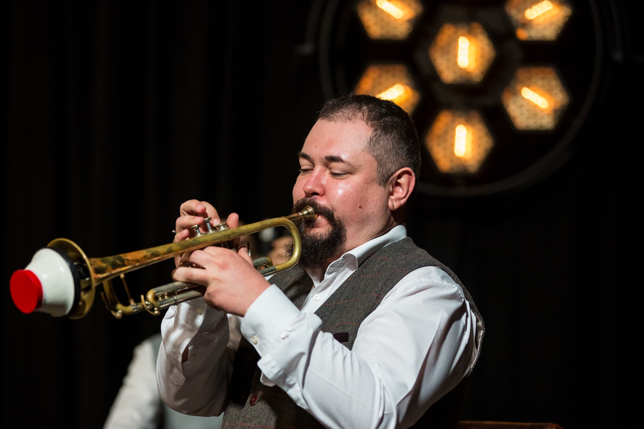 Man playing trumpet in a dark room with decorative lighting in the background.