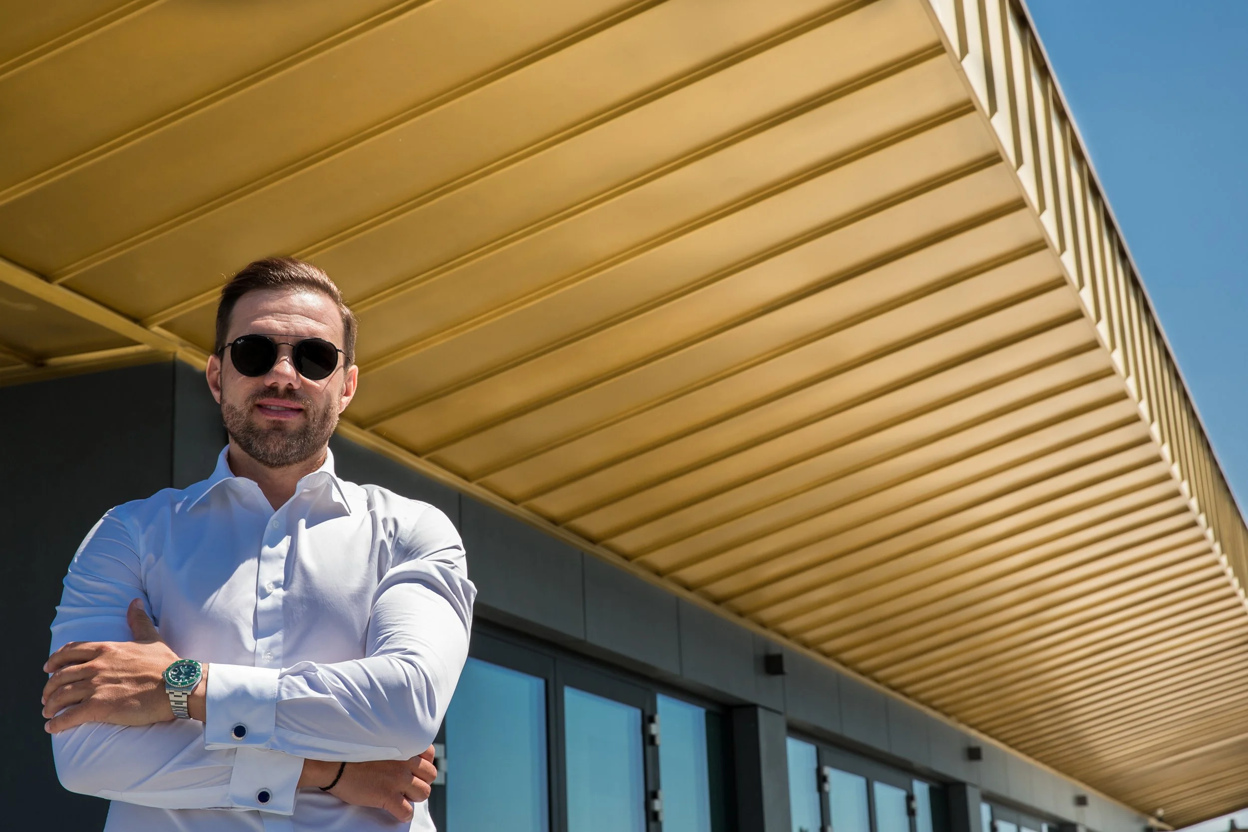 A man wearing sunglasses and a white shirt with rolled-up sleeves standing with arms crossed in front of a modern building with a yellow overhang and blue sky.