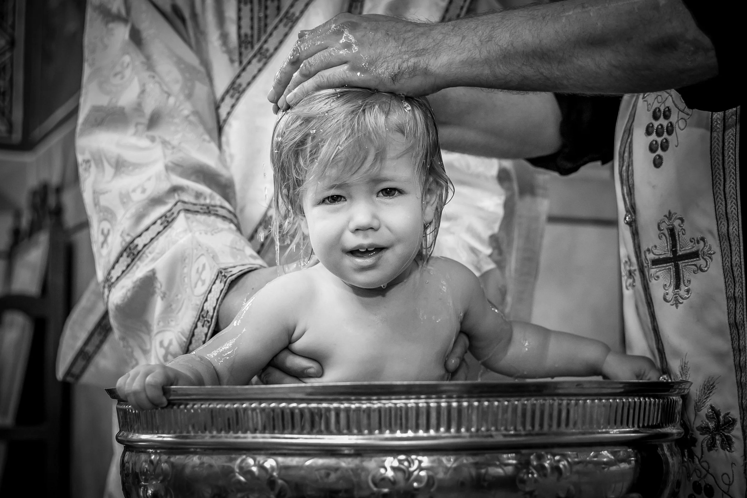 A young child having a religious water baptism, with an adult hand on the child's head, in a church setting with religious cloth and objects visible.