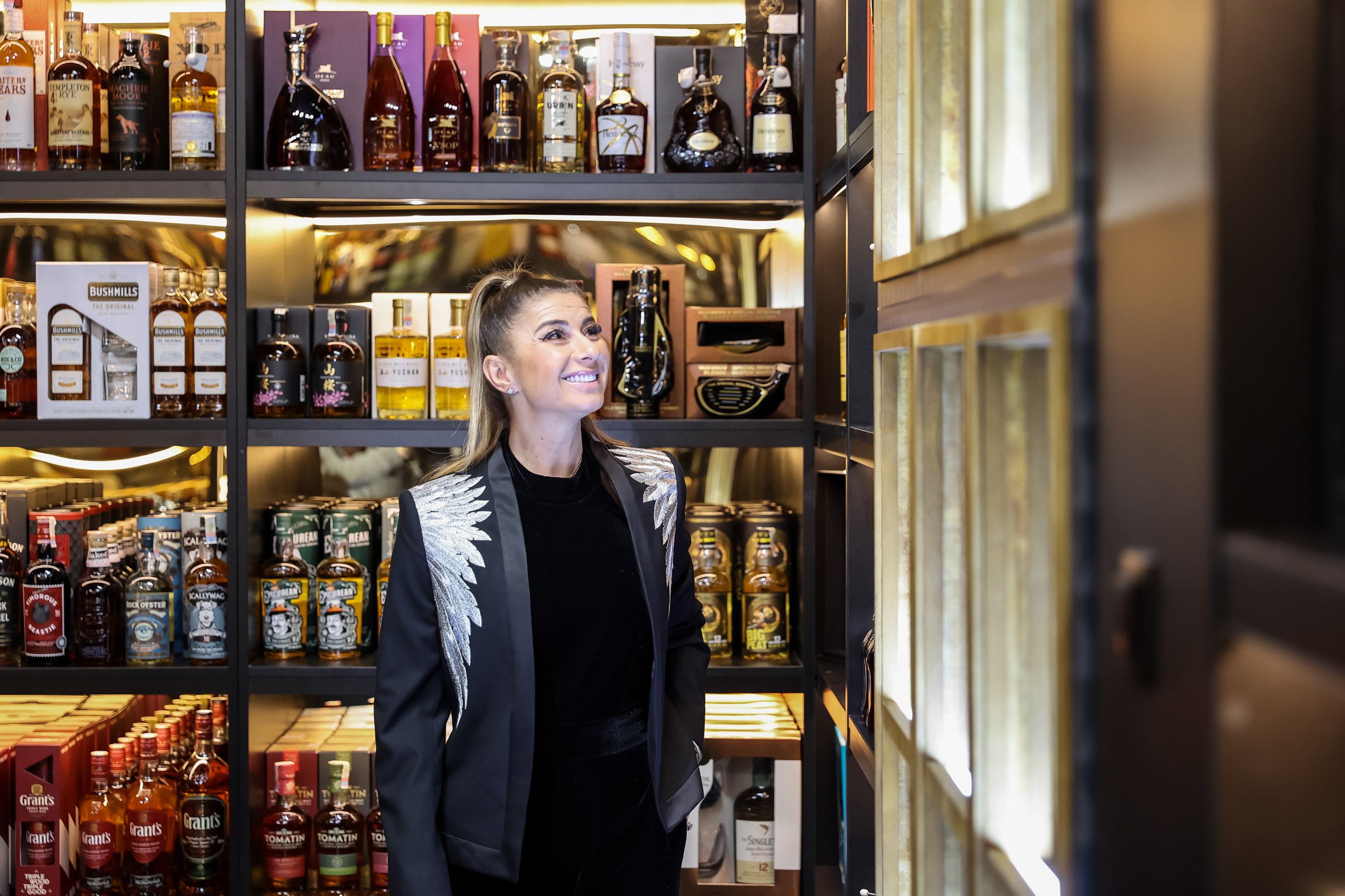 A woman with long hair styled in a ponytail and wearing a black blazer with silver embroidery on the shoulders, smiling while shopping for liquor in a store with shelves stocked with various bottles of whiskey and other spirits.