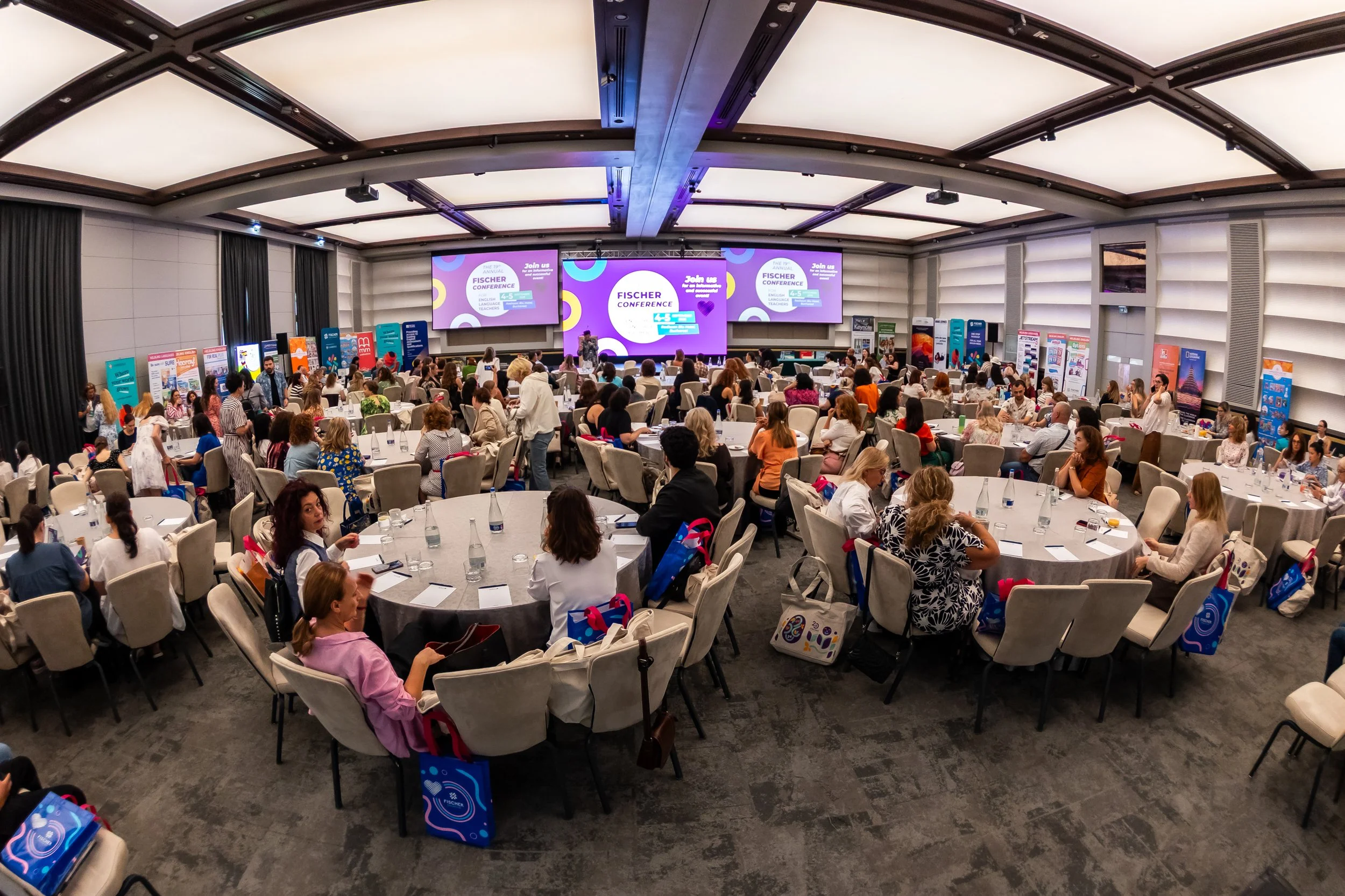 A large conference room filled with round tables and chairs, where many people are attending a conference. There is a big screen at the front displaying the Fischer Conference logo, and several banners lined along the walls.