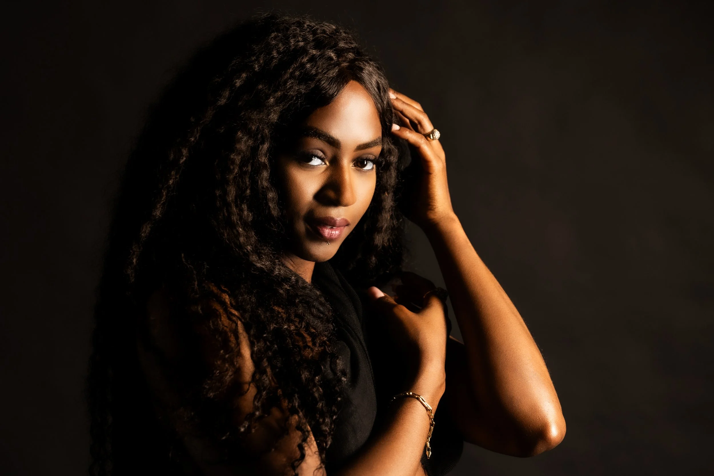 portrait of a woman with long curly hair posing with her hand on her head against a dark background