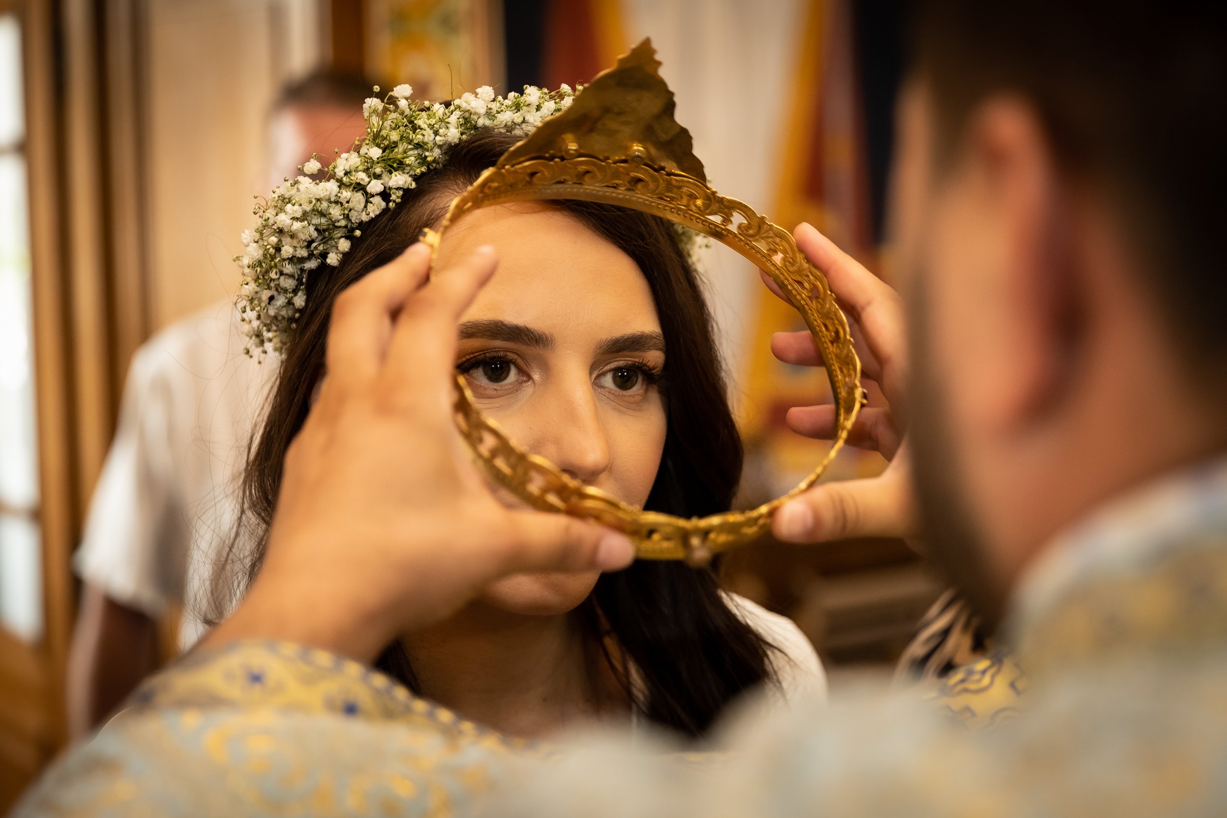 A woman with dark hair and brown eyes wearing a floral crown is looking at herself in a mirror while someone adjusts a gold headpiece on her head.
