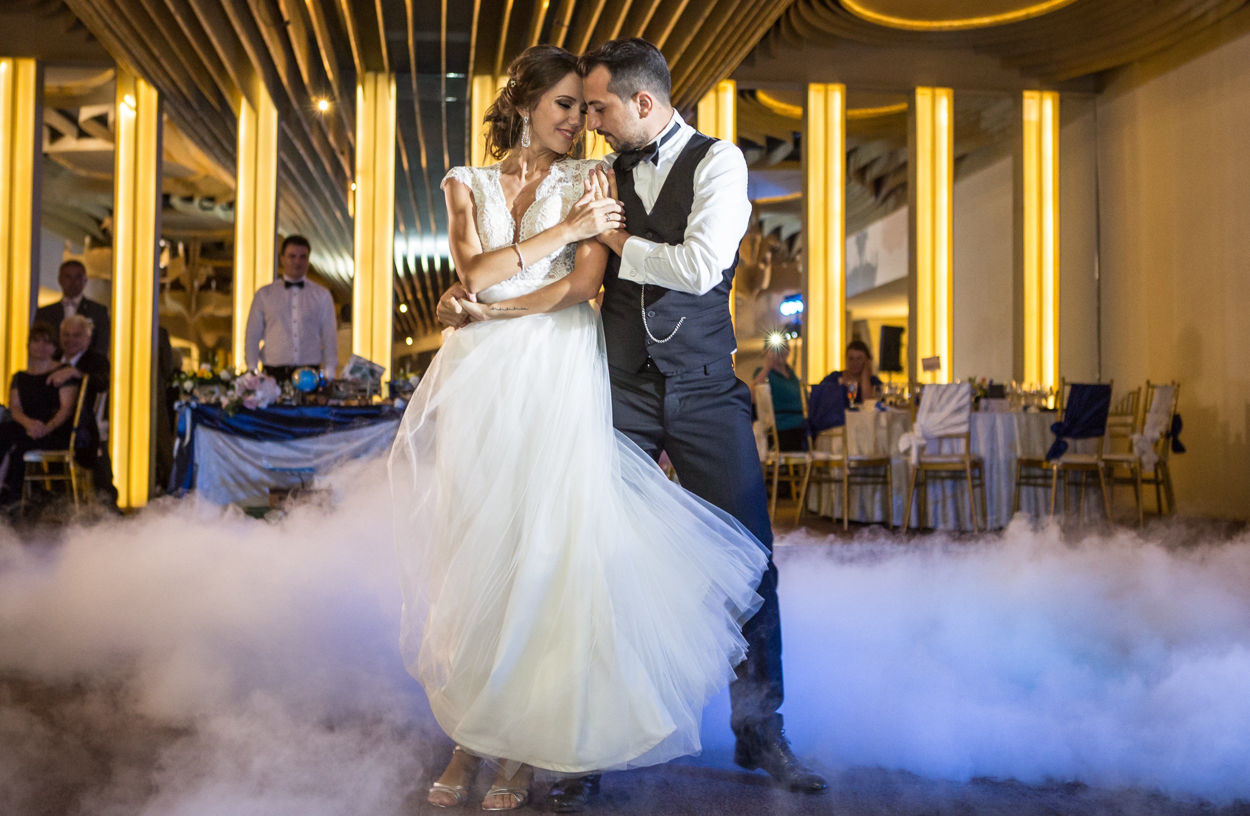 A bride and groom sharing a dance at their wedding reception, surrounded by guests and illuminated by warm lighting with modern decor.