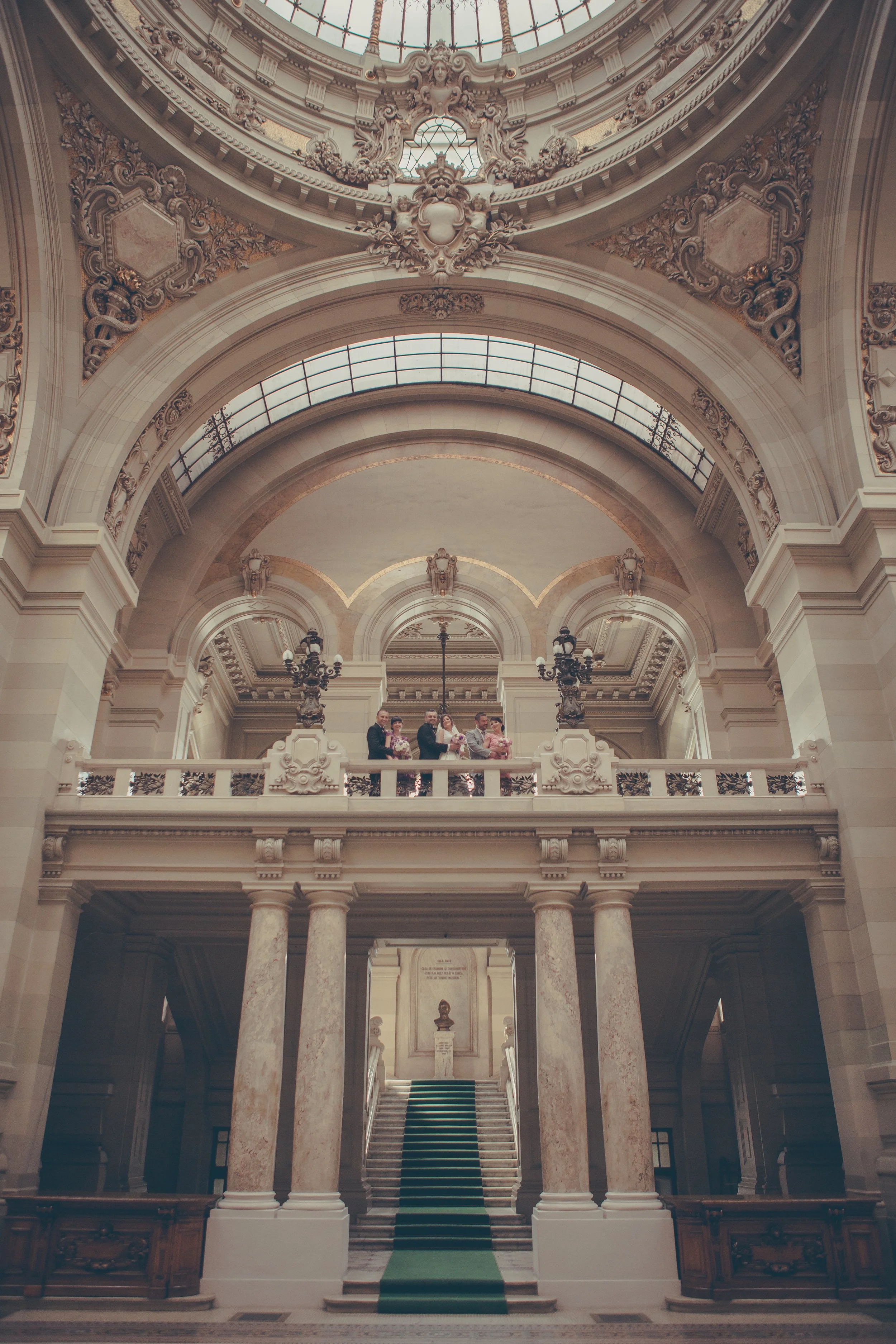 Interior view of a grand ornate building with a staircase, marble columns, and elaborate architectural details like arches and sculptures, with a group of people standing on a balcony.