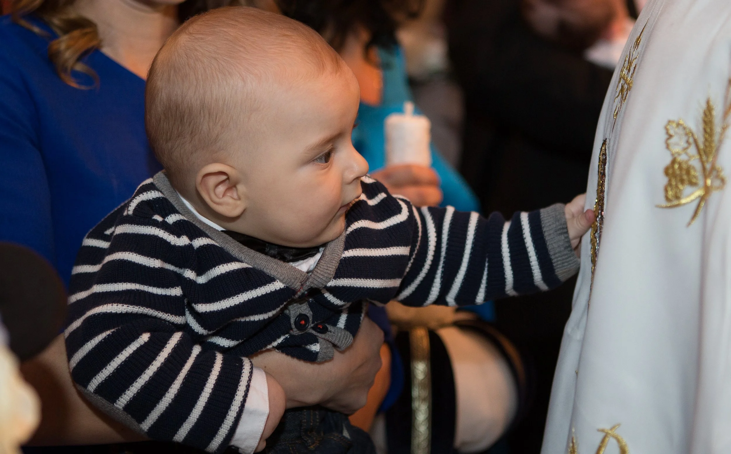 A young boy with light skin and short blond hair, wearing a navy and white striped sweater, is reaching out to touch an ornate, embroidered religious vestment while sitting on someone's lap in a crowded indoor setting.