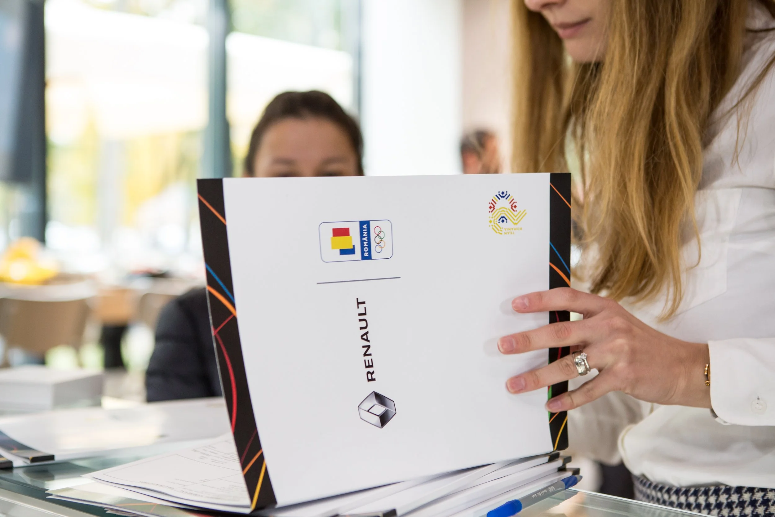 Close-up of a woman holding a Romania Olympic incentives booklet, with a woman in the background at a desk with papers and pens.