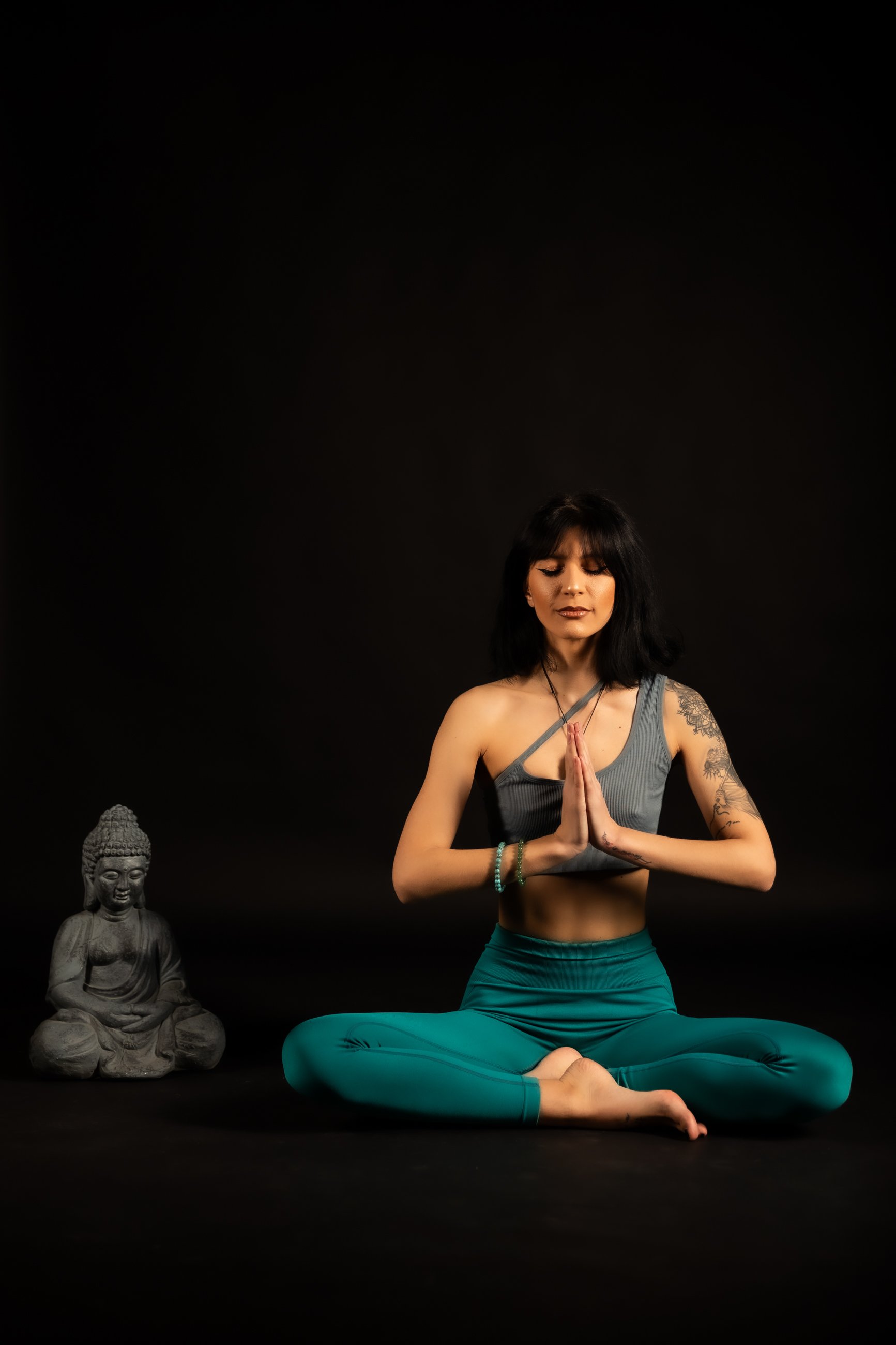 Woman practicing yoga in a cross-legged seated posture with hands in prayer position, wearing yoga attire, tattoos on arms, with a small Buddha statue beside her against black background.