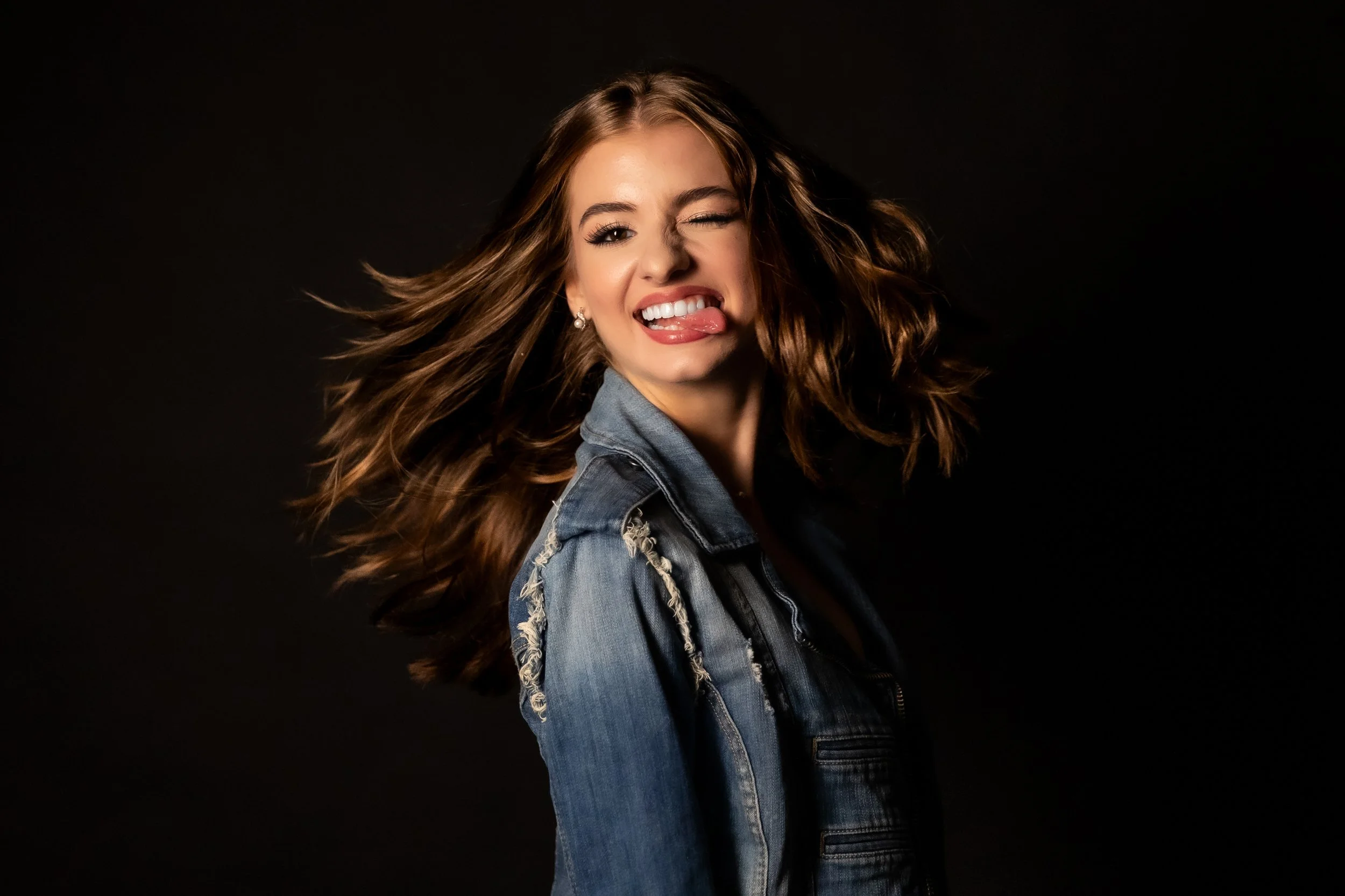A young woman with long wavy hair, wearing a denim jacket, sticking her tongue out and winking against a dark background.