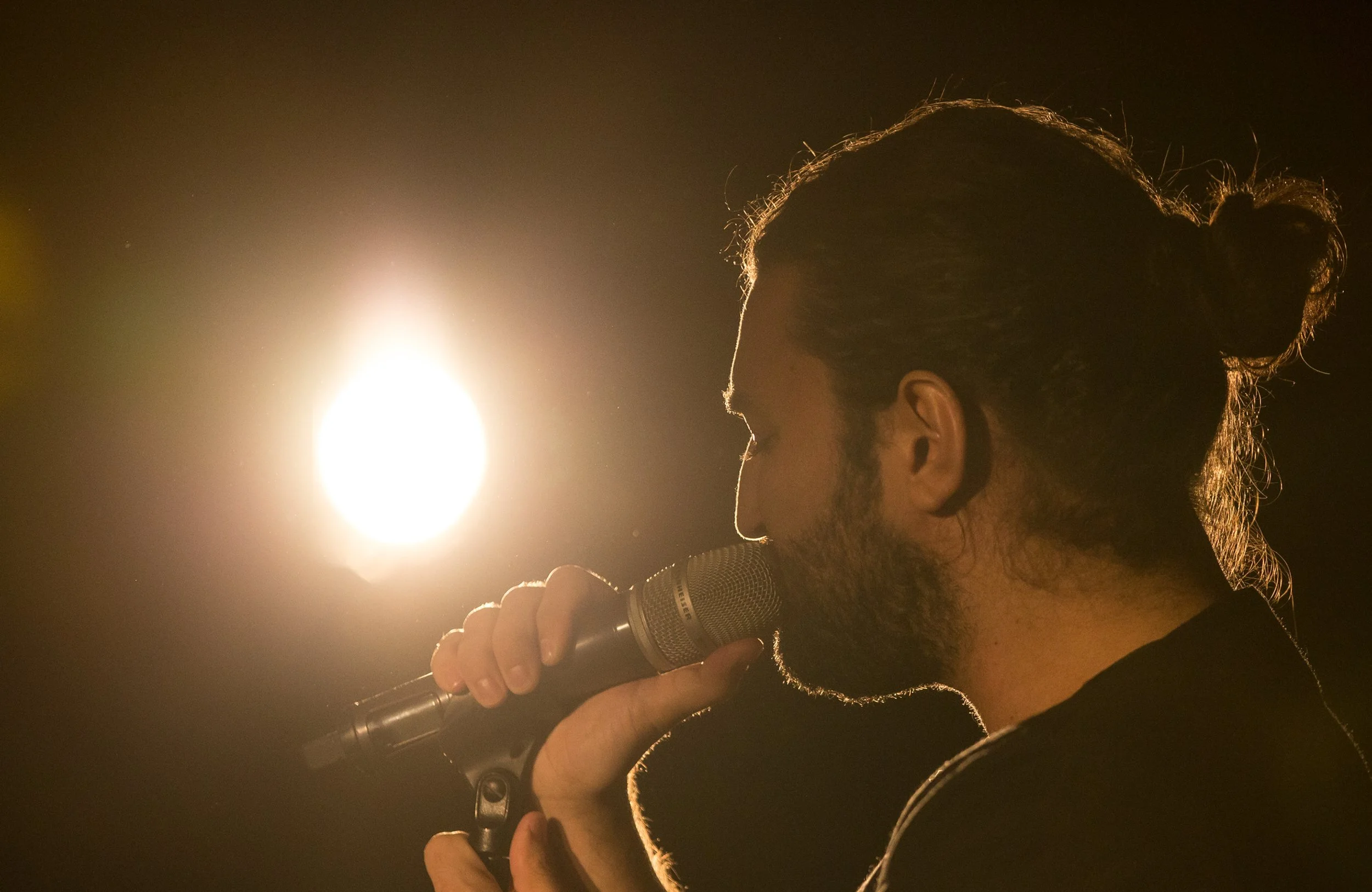 A person with a bun hairstyle singing into a microphone in front of a bright light.