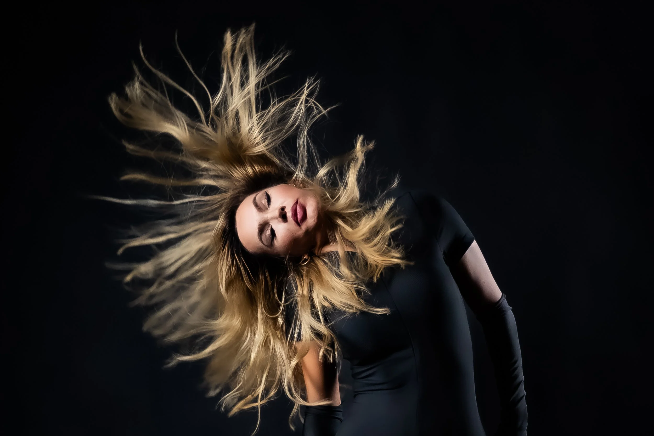A woman with long blonde hair wearing a black outfit, with her hair flowing and her eyes closed, standing against a black background.