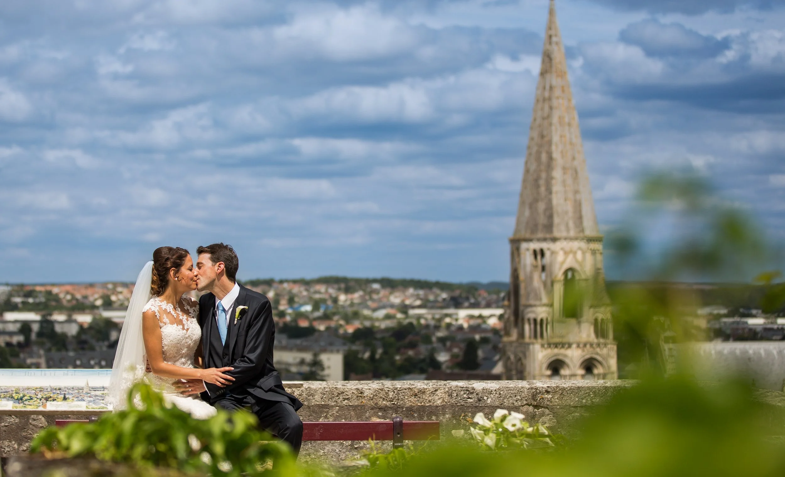 A bride and groom kissing on a balcony with a view of a town and a large church with a tall spire in the background under a cloudy sky.