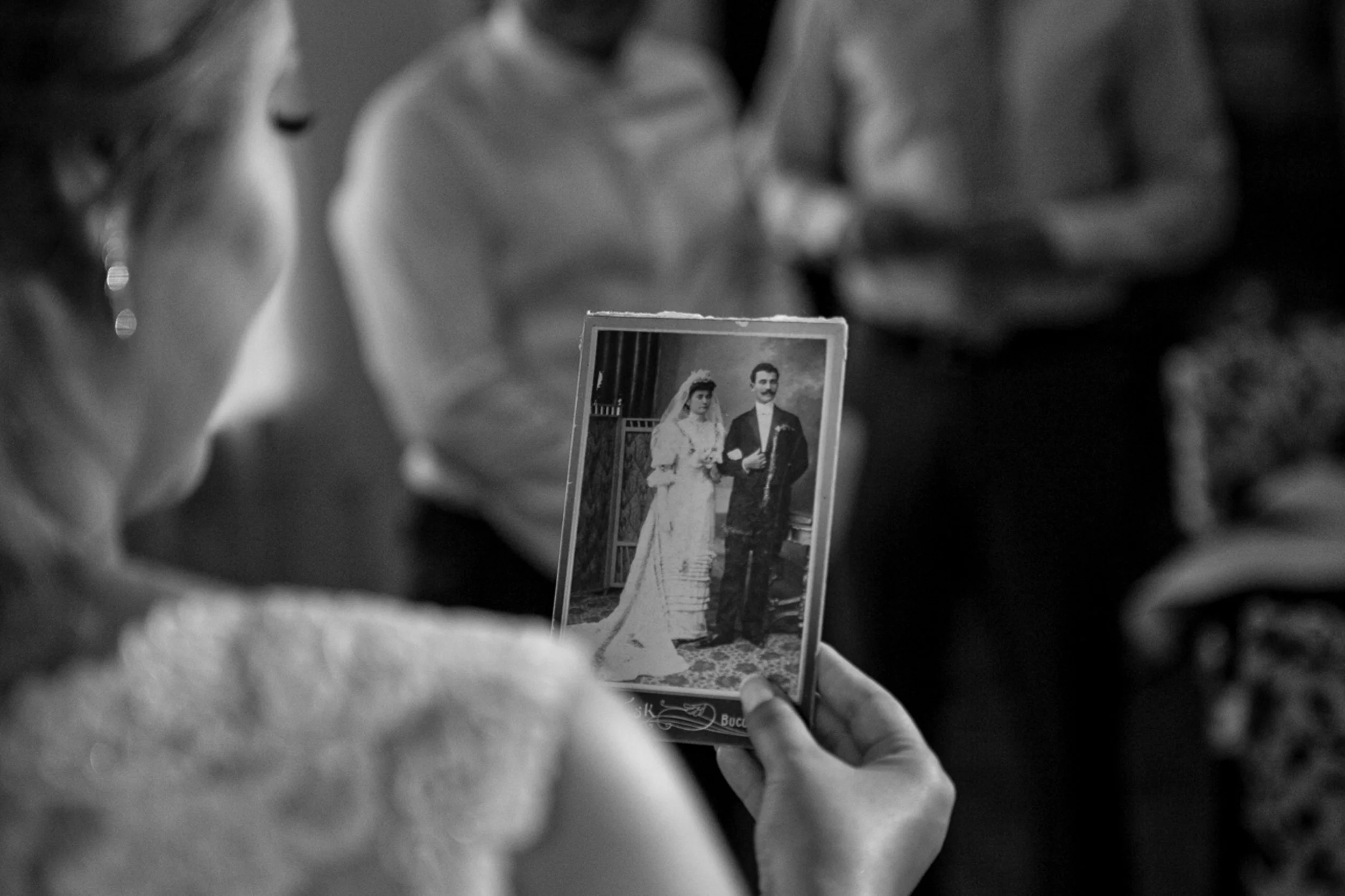 A person holding a photograph of a wedding couple, with another person visible in the background, in a black and white photo.