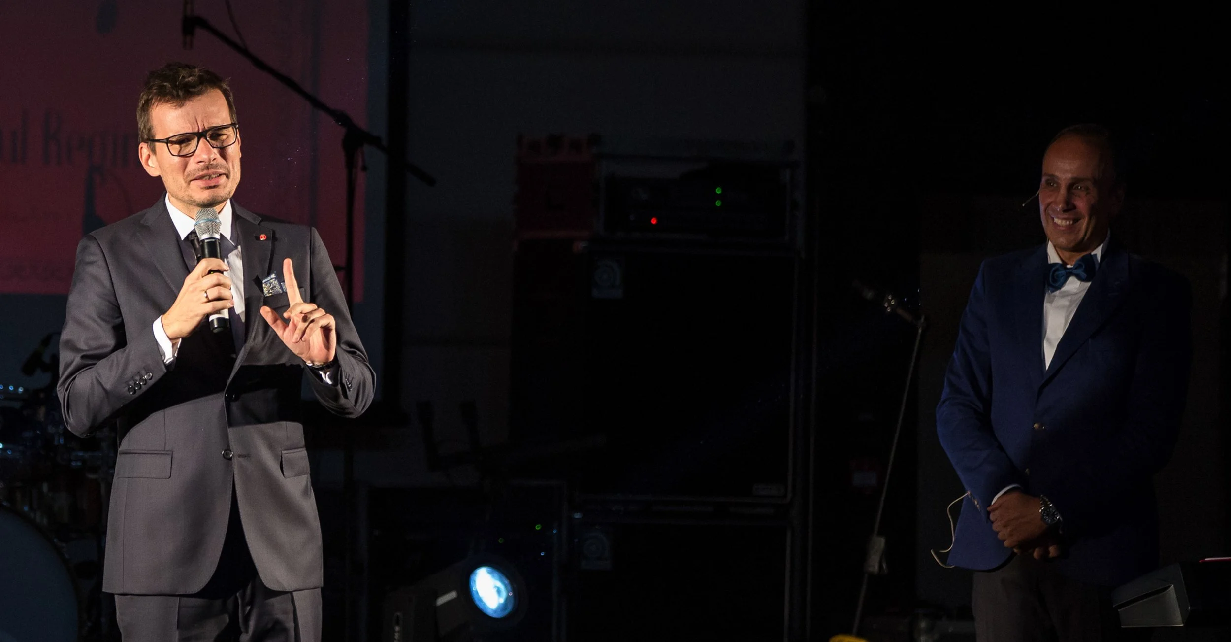 A man in a dark suit with glasses and a microphone appears to be speaking or singing with his eyes closed. Another man in a dark suit with a bow tie stands smiling in the background. The setting is dark, possibly on a stage, with some equipment visib