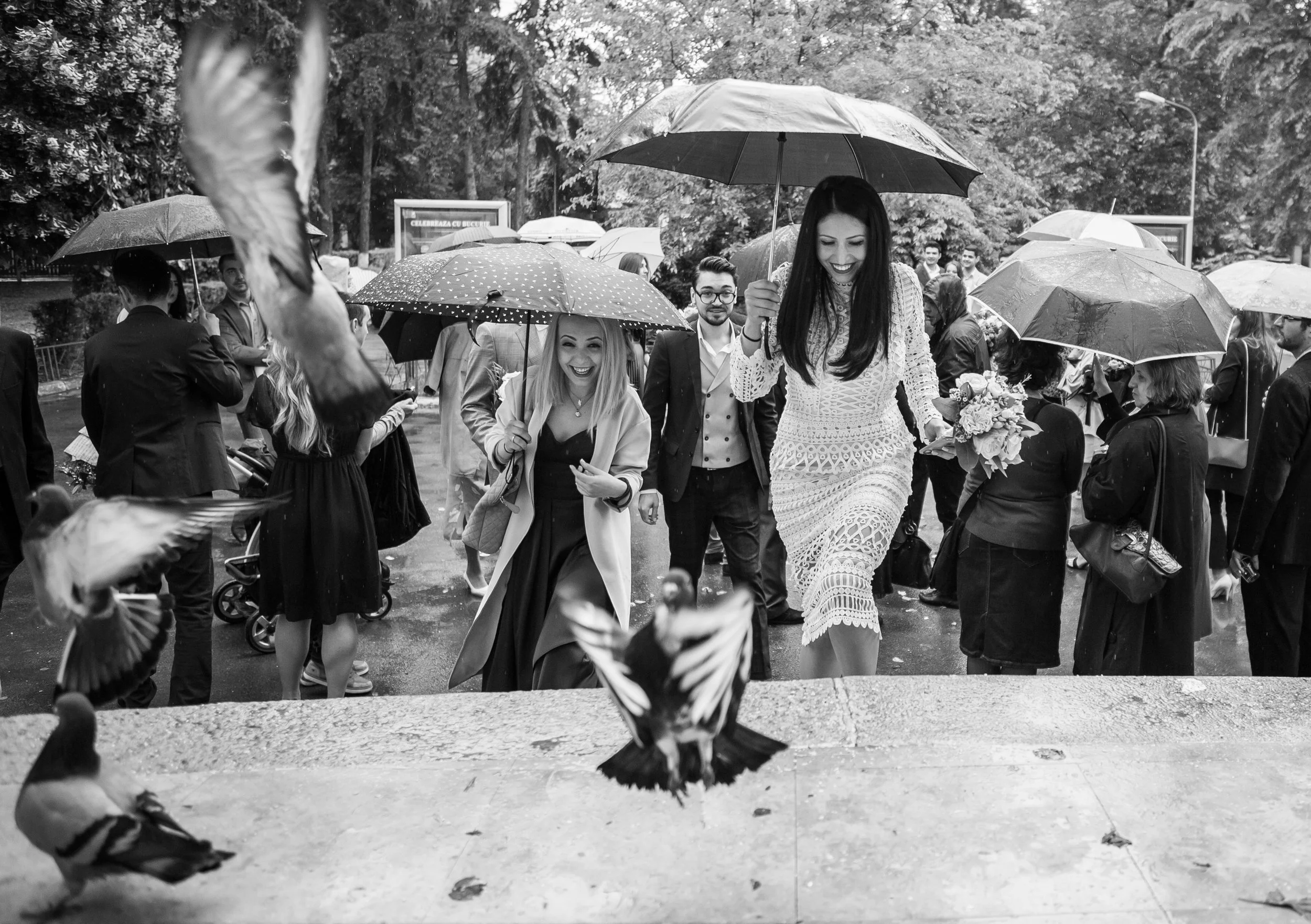 People walking in the rain holding umbrellas, with some smiling and enjoying the moment, in a park or outdoor area with trees.