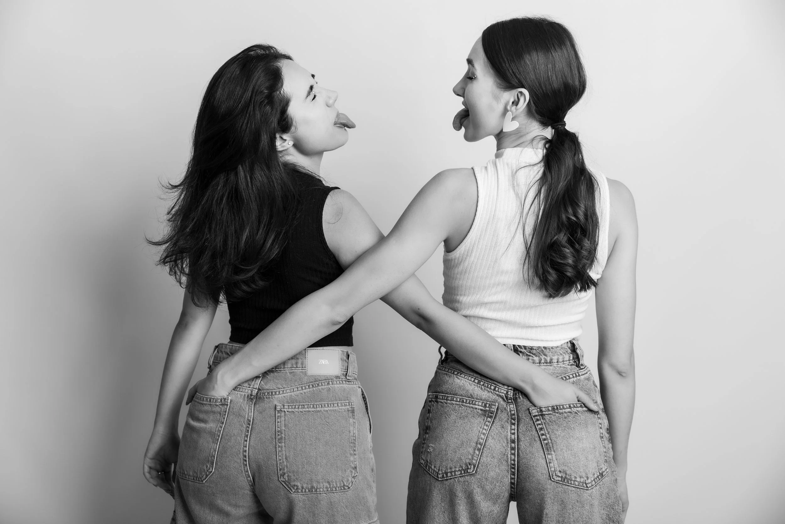 Two young women facing each other with their tongues out, one wearing a black top and the other a white top, both in jeans, standing against a plain background.