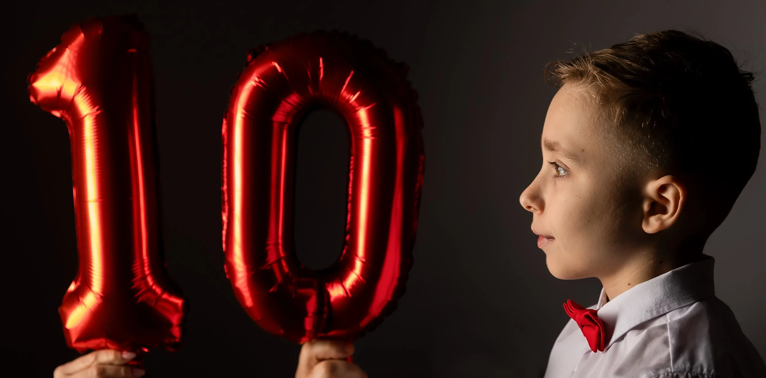 A young boy dressed in a white shirt with a red bow tie, holding red foil balloons in the shape of the number '10', looking at them against a dark background.