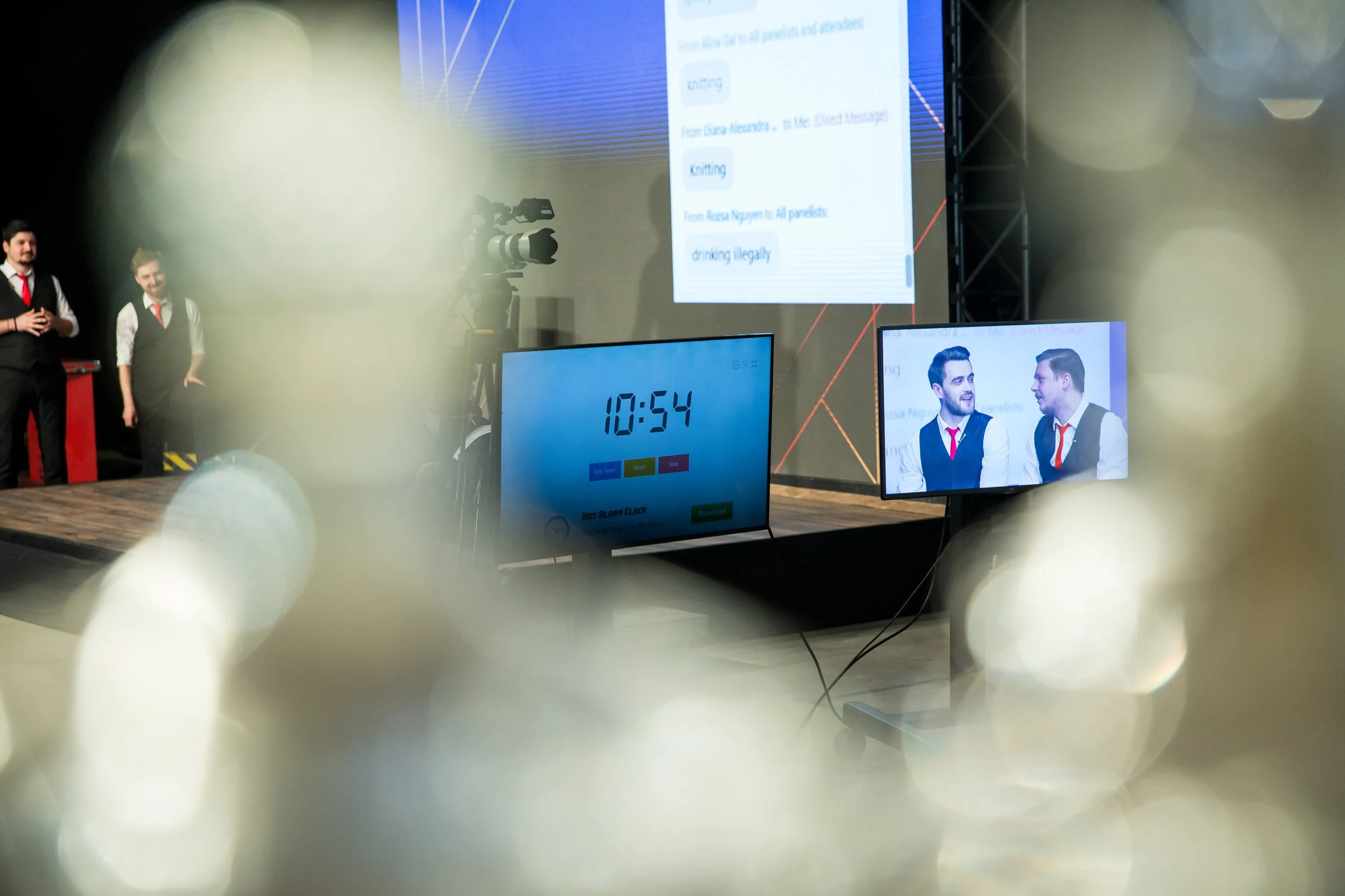 Two men in suits and ties standing on stage at a conference or presentation, with a large screen behind them displaying text about knitting and illegal drinking. There are multiple monitors in front showing a timer and a photo of the two men, and a c