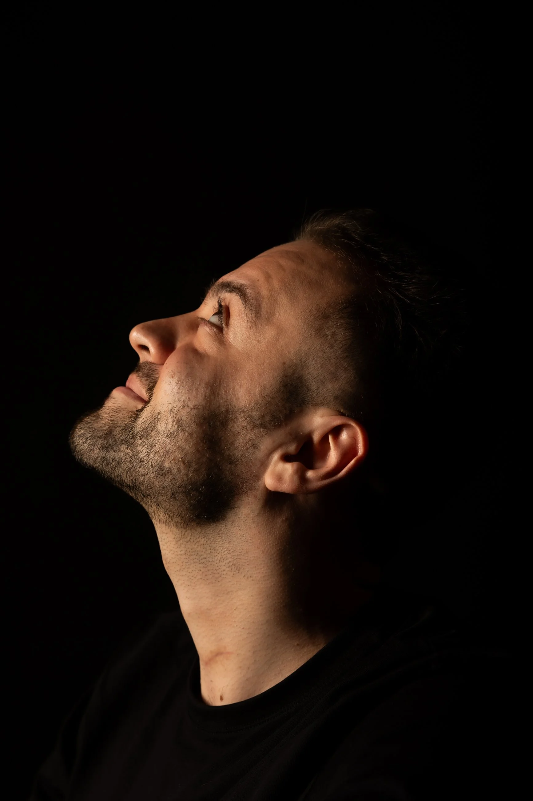 Close-up side profile of a man with short dark hair and a beard, looking upward against a black background.