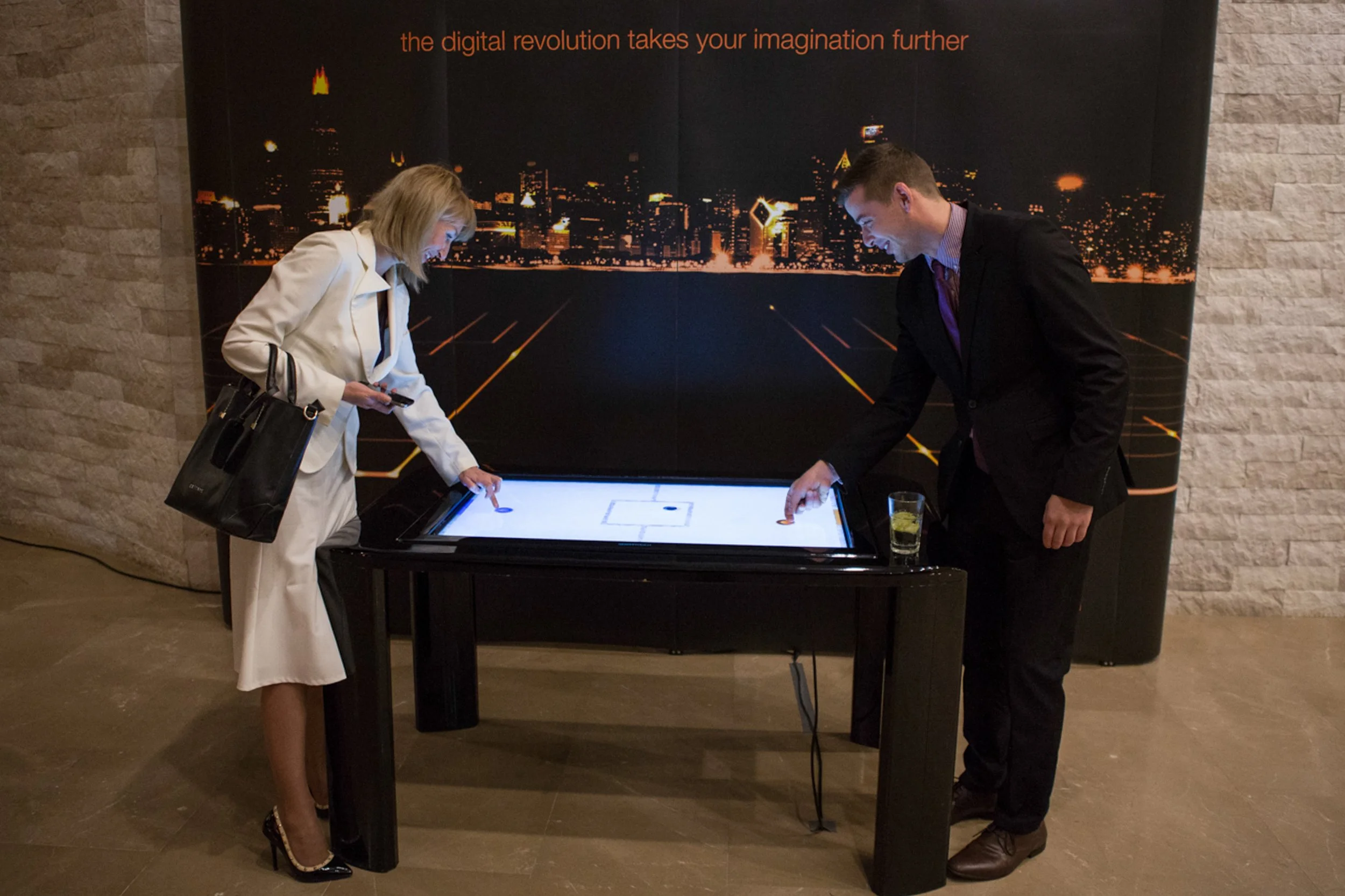 Two people, a woman and a man, are playing air hockey on a digital table. The woman is wearing a white blazer and black shoes, and the man is in a black suit. They are smiling and leaning over the table, hitting the puck with paddles. The background 