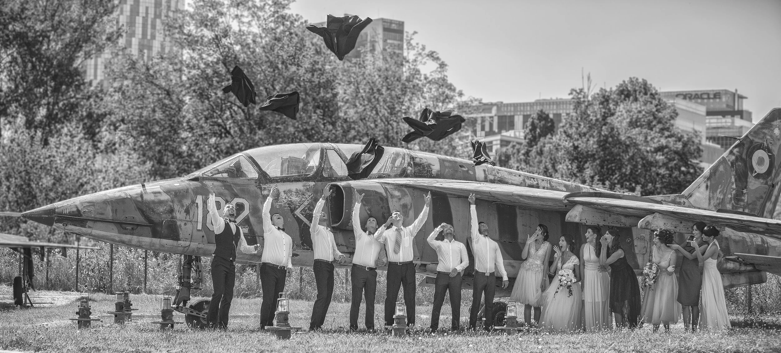 A group of people, including men and women in formal and wedding attire, celebrating in front of a military jet plane during a wedding or special event. Some are tossing jackets into the air, with wedding dresses and bouquets visible among the group.