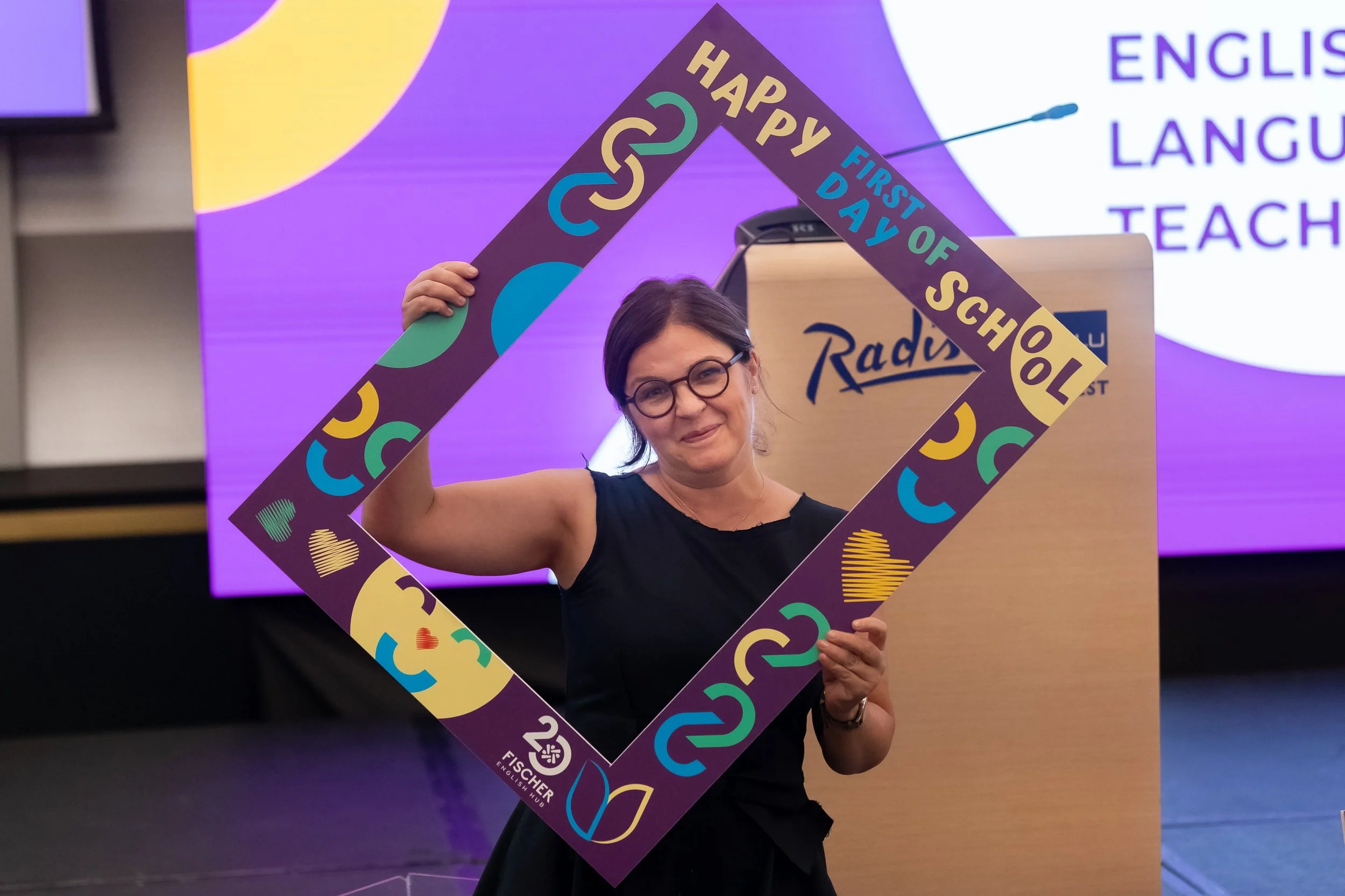 A woman wearing glasses holds a colorful frame with the words "Happy First Day of School" at a school event or celebration.