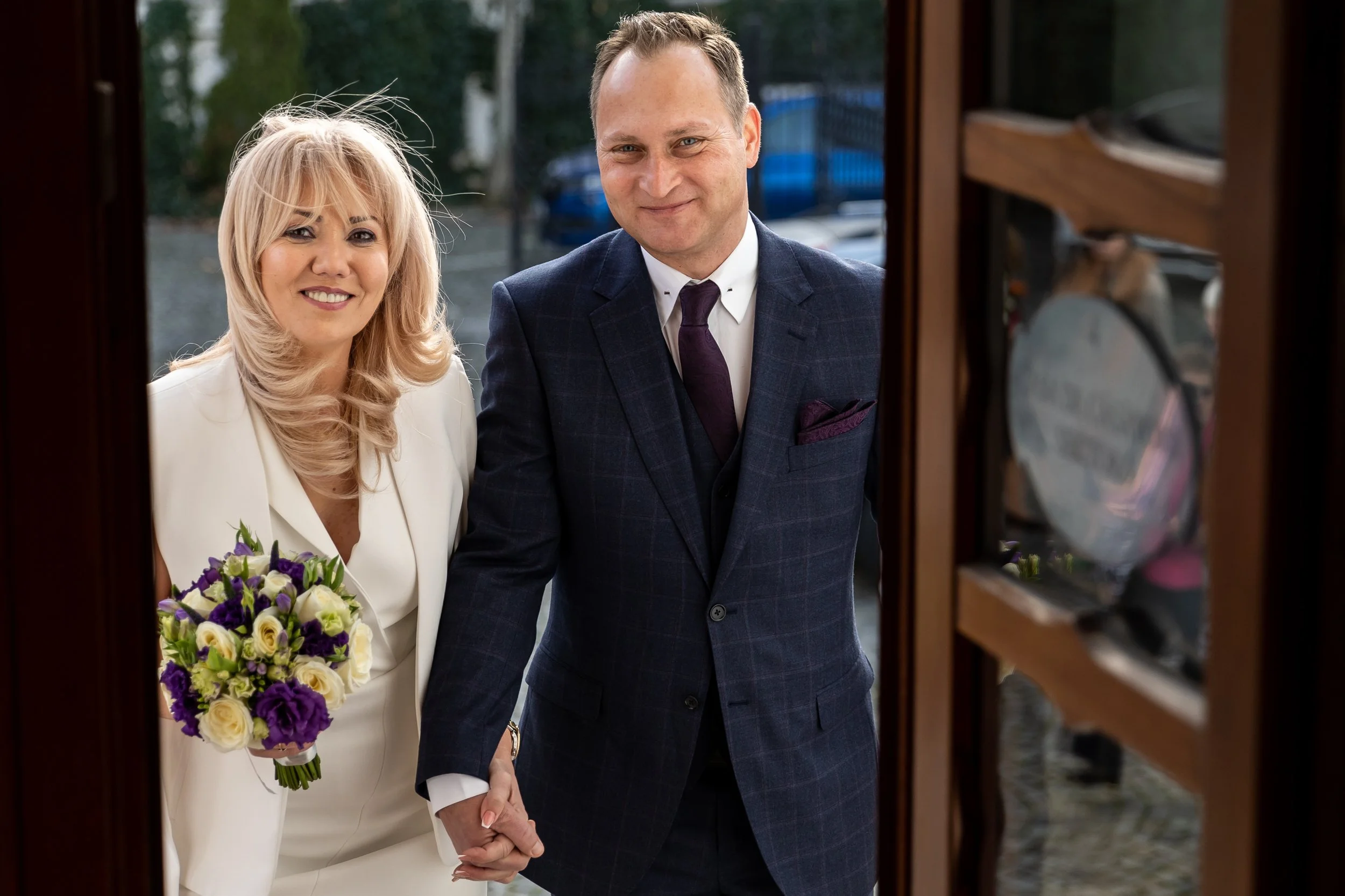 A smiling bride and groom holding hands outside a building with a wooden door, the bride holding a bouquet of purple and white flowers, dressed in a white gown, and the groom in a dark checkered suit with a white shirt and tie.
