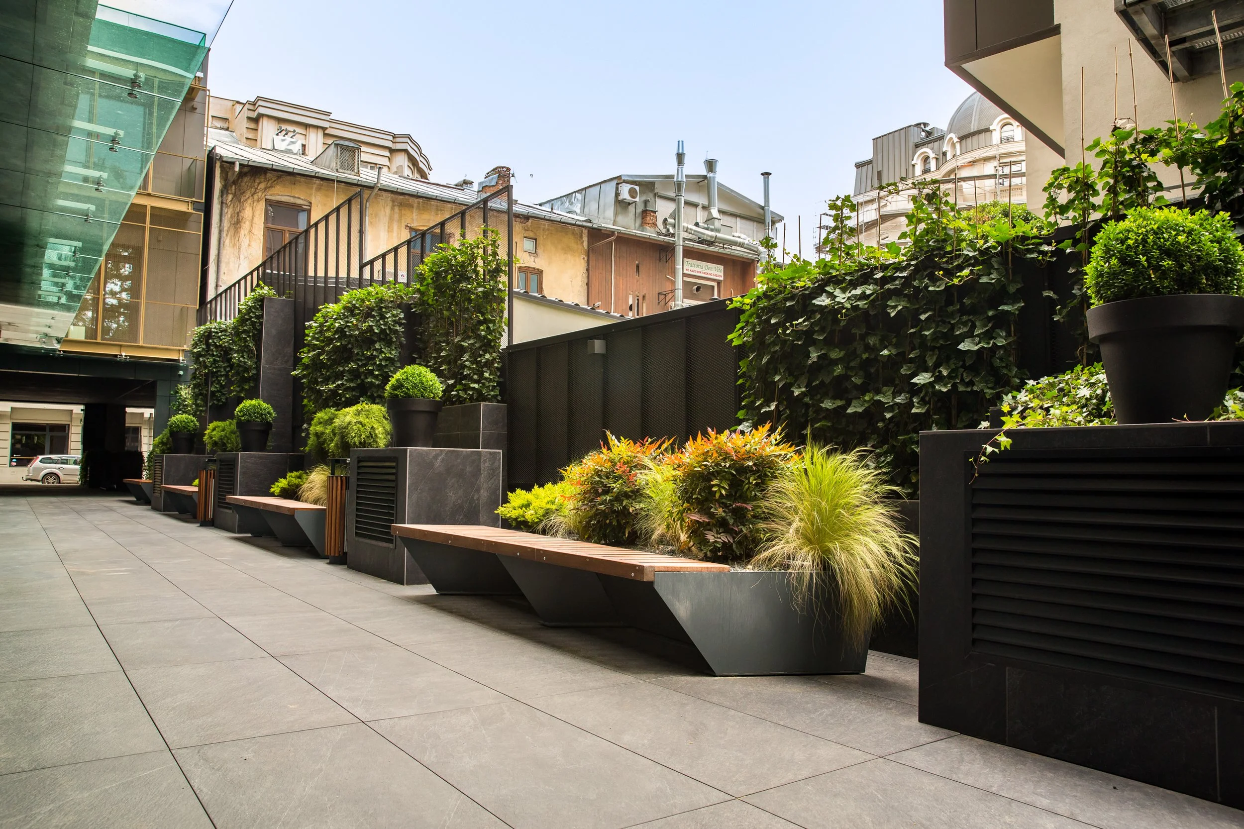 Urban outdoor space with potted plants, benches, and modern decor, surrounded by older and newer buildings under a clear sky.