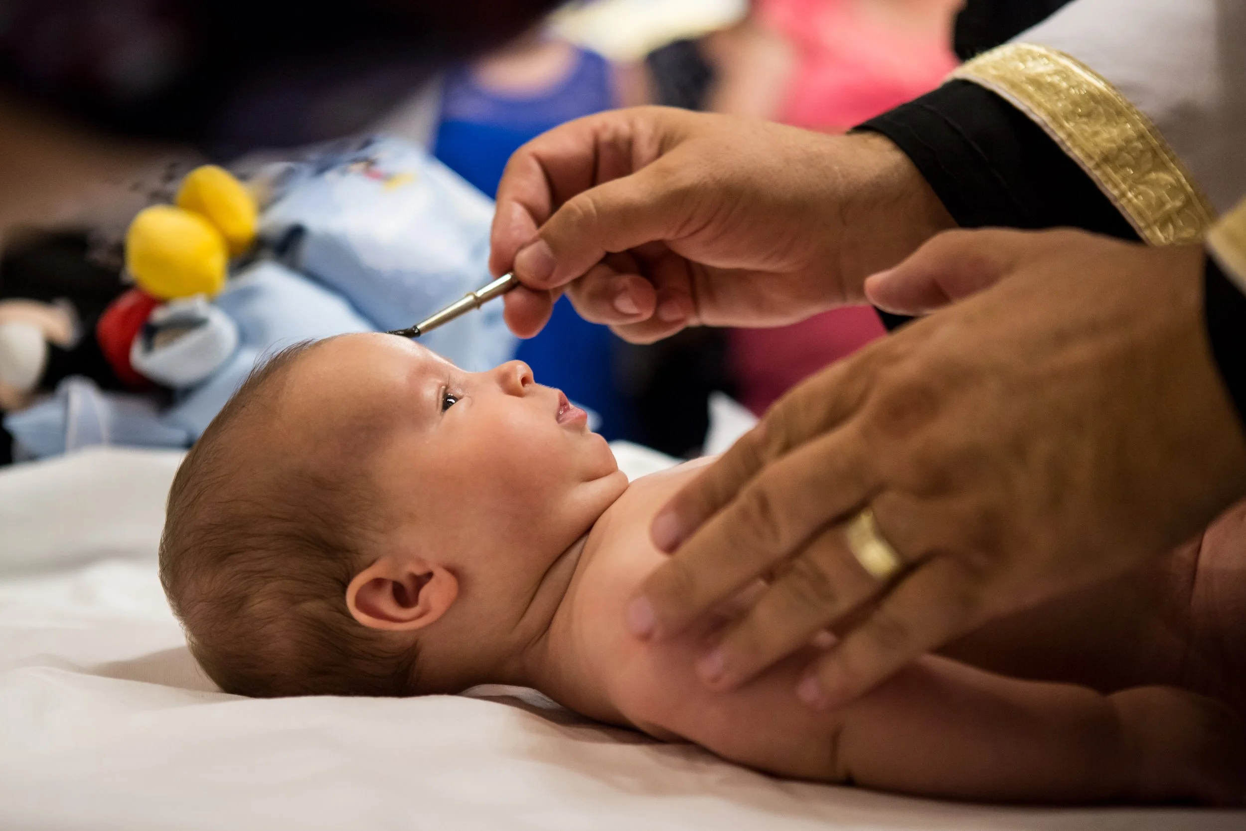 Close-up of a newborn baby lying on a hospital bed, receiving a vaccination with a healthcare professional's hand holding a syringe gently administered to the baby's upper arm, with medical equipment and blurred people in the background.