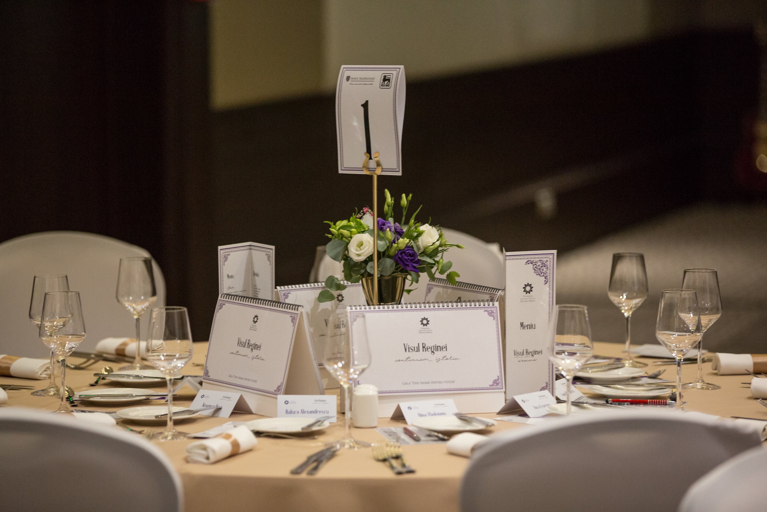 A table set for a formal event with white chairs, a floral centerpiece, wine glasses, and place cards, and a table number sign with the number 1.