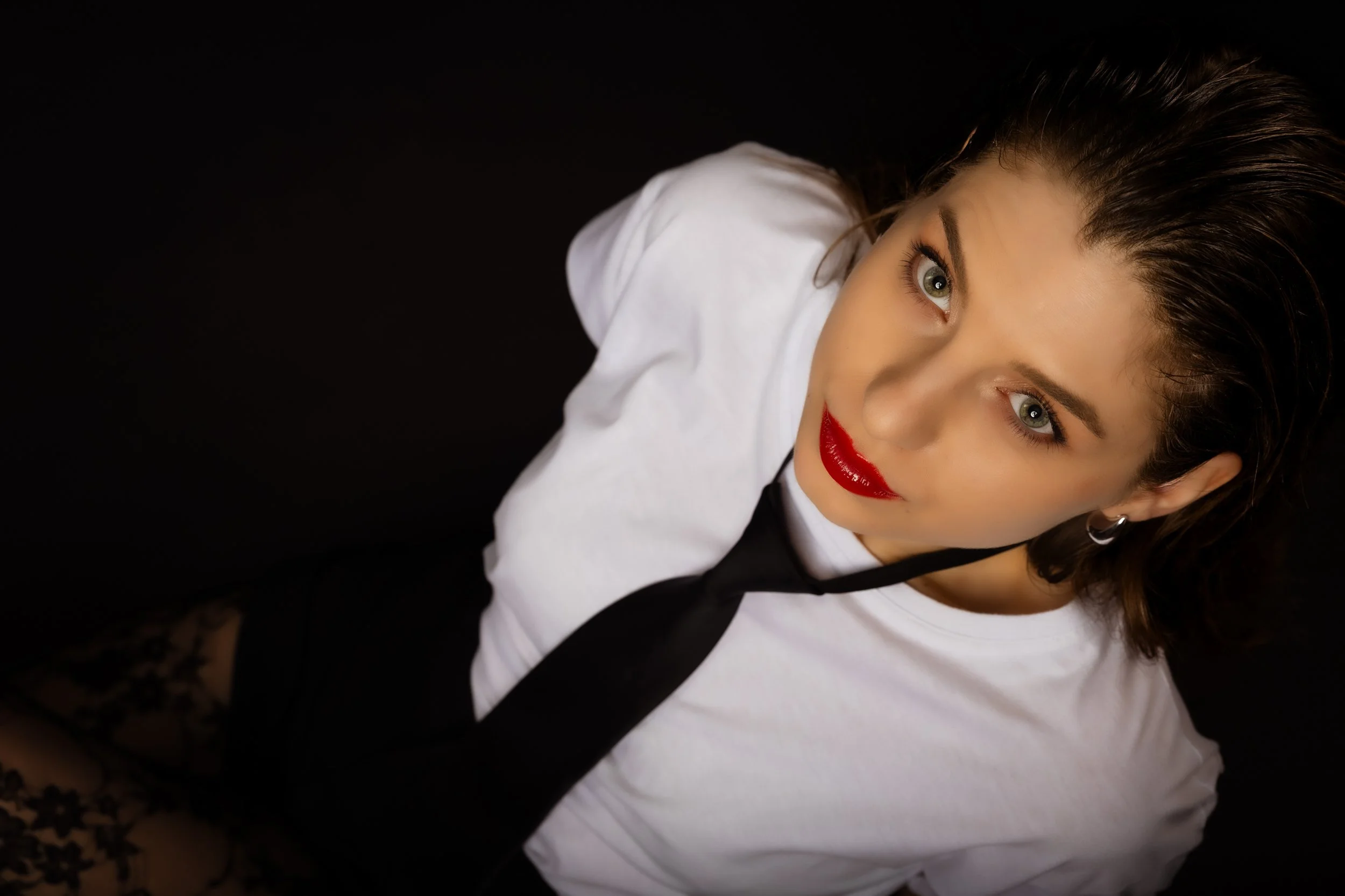 A woman with dark hair, wearing red lipstick, hoop earrings, a white t-shirt, and a black tie, looking at the camera against a black background.