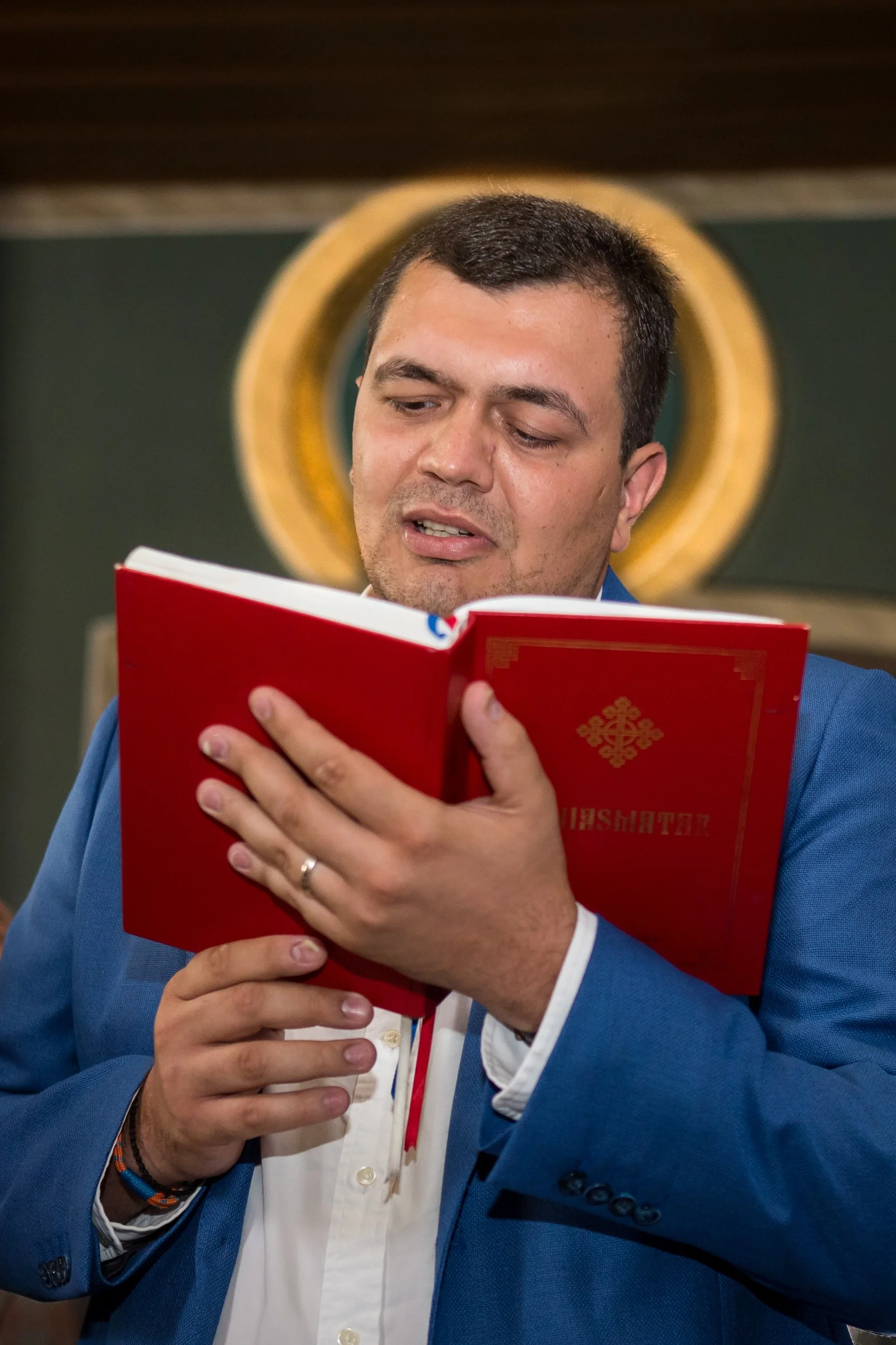 A man in a blue suit and white shirt holding a red book with gold design, reading intently, with a presidential seal in the background.