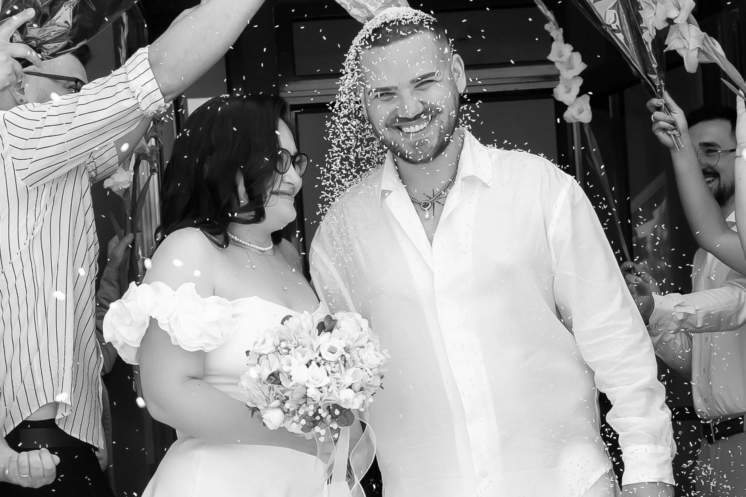 A smiling man and woman at a wedding celebration, surrounded by confetti and flowers, with several people holding flower arrangements around them.